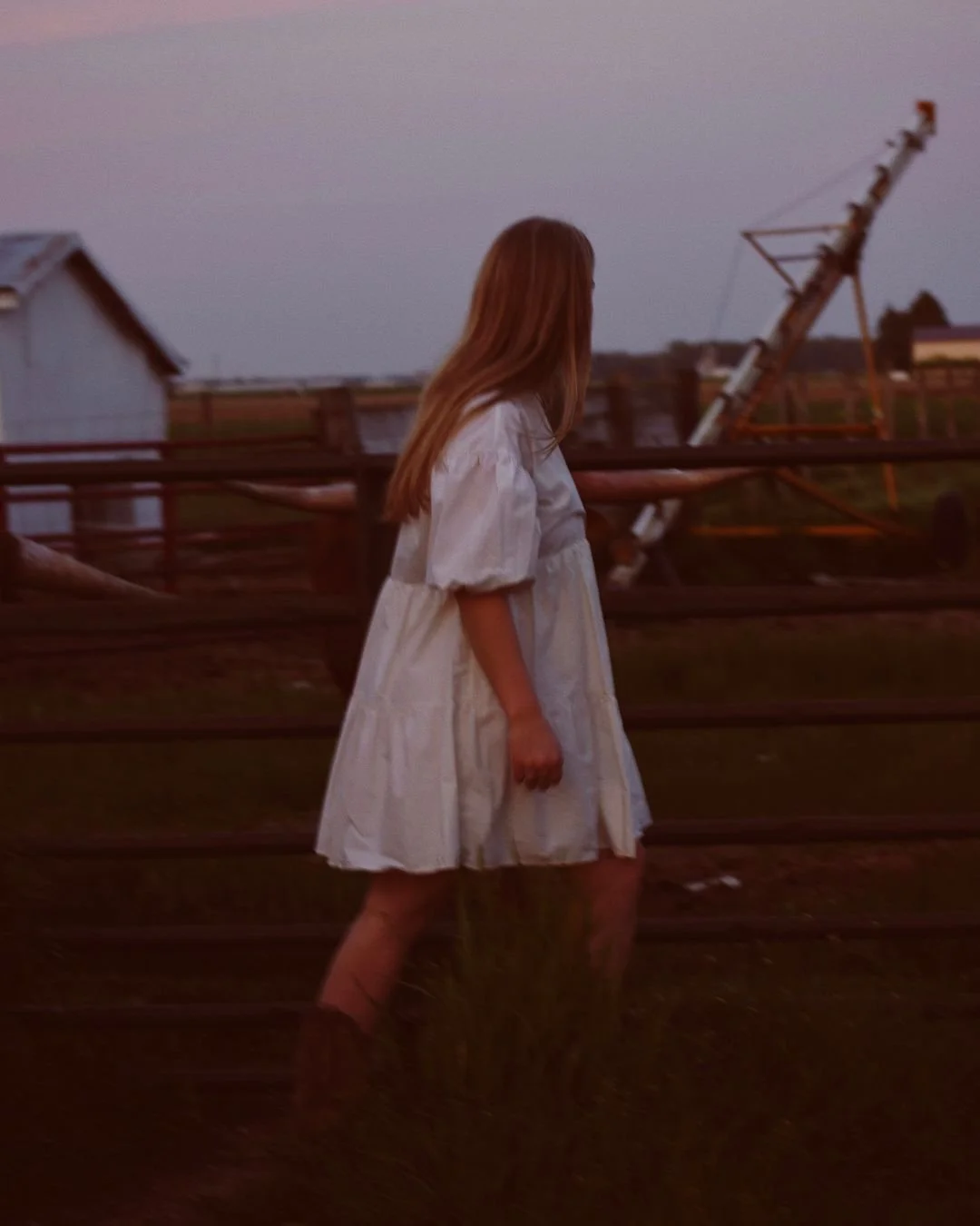 A girl in a white dress walking near railroad tracks at dusk, with damaged structures and a tilted utility pole in the background.
