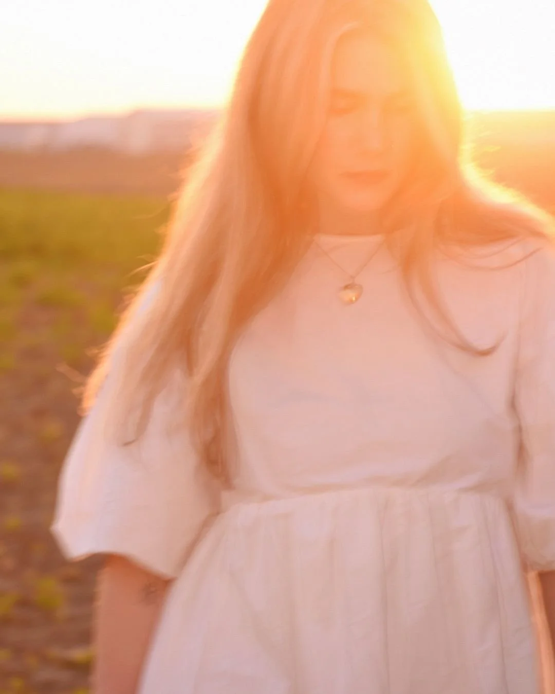 A woman with long hair wearing a white dress and a heart-shaped necklace standing outdoors during sunset.