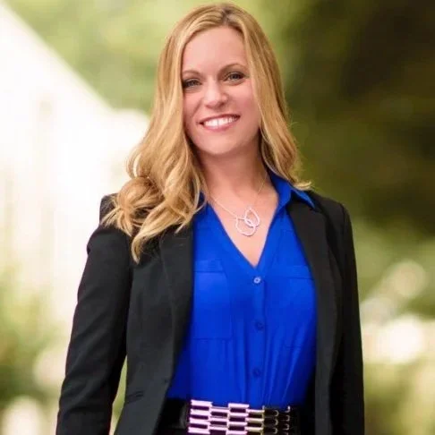 A smiling woman with glasses and light brown wavy hair, wearing a black shirt, standing in front of a green plant wall.