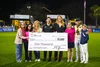 A group of people standing on a sports field at night, holding a large check for $1,000 made out to 'Leveling The Fields' from The Ideal Life, with the memo reading “Community hero.”