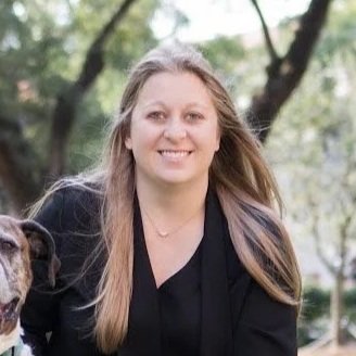 A blonde woman wearing a black shirt and green pants poses outdoors beside a brown and white dog.
