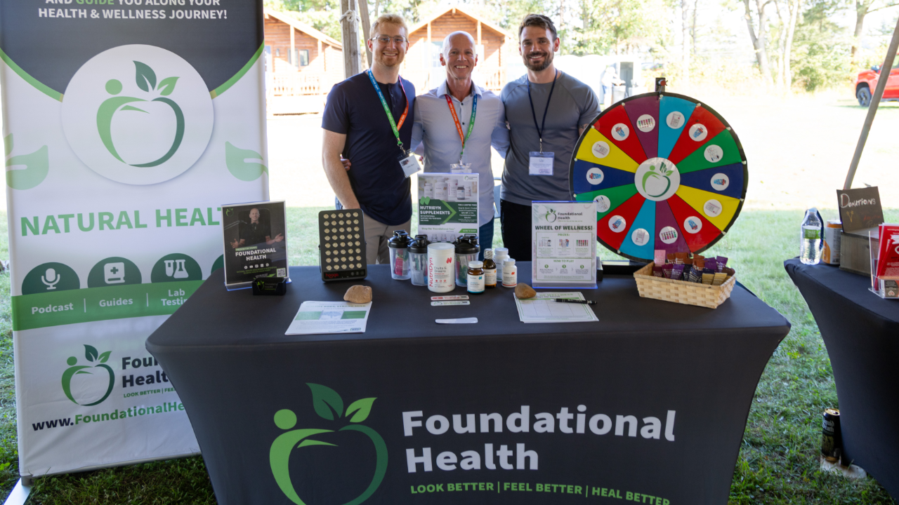 Three men stand behind a promotional table for Foundational Health at an outdoor event, with a colorful wheel of wellness and various health supplements on display.