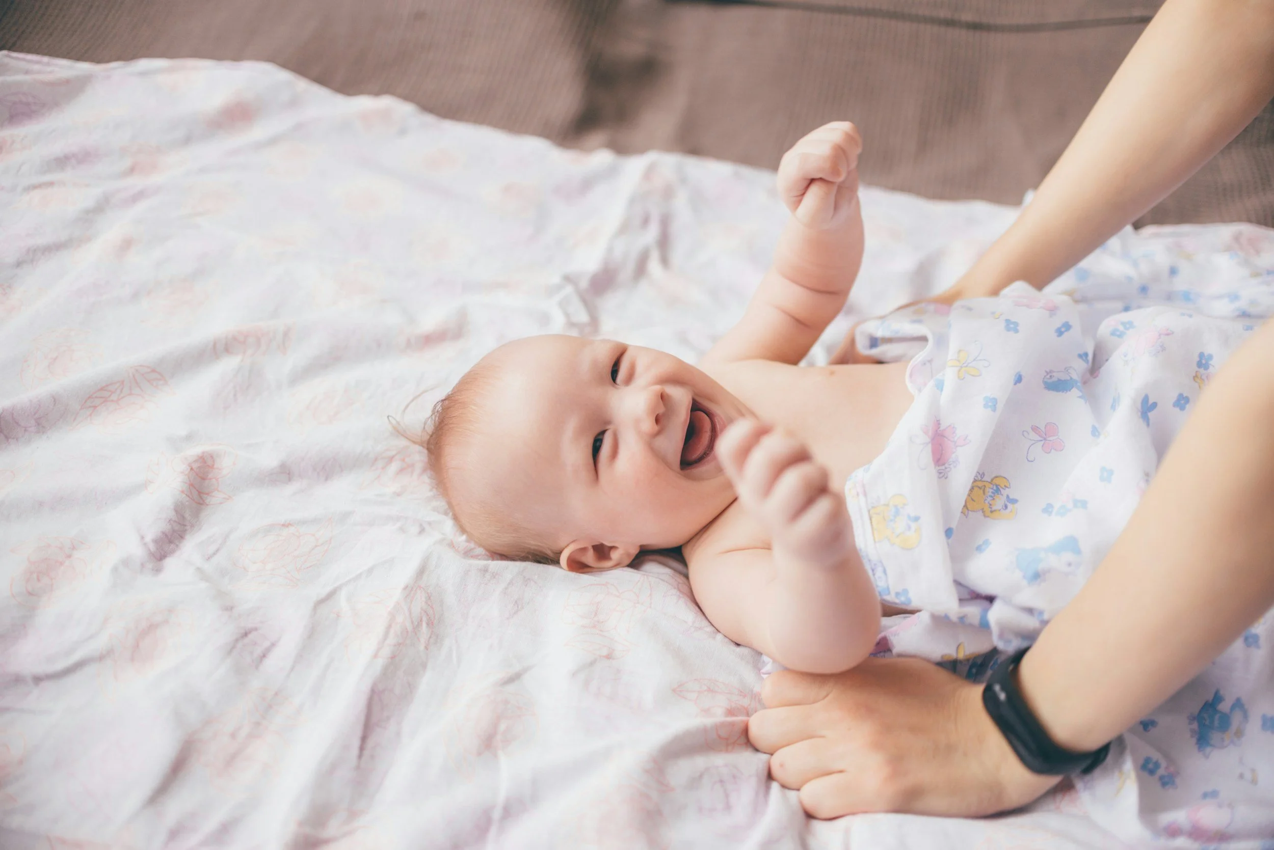 A smiling baby lying on a blanket, being held by an adult's hands, with a look of joy and excitement.
