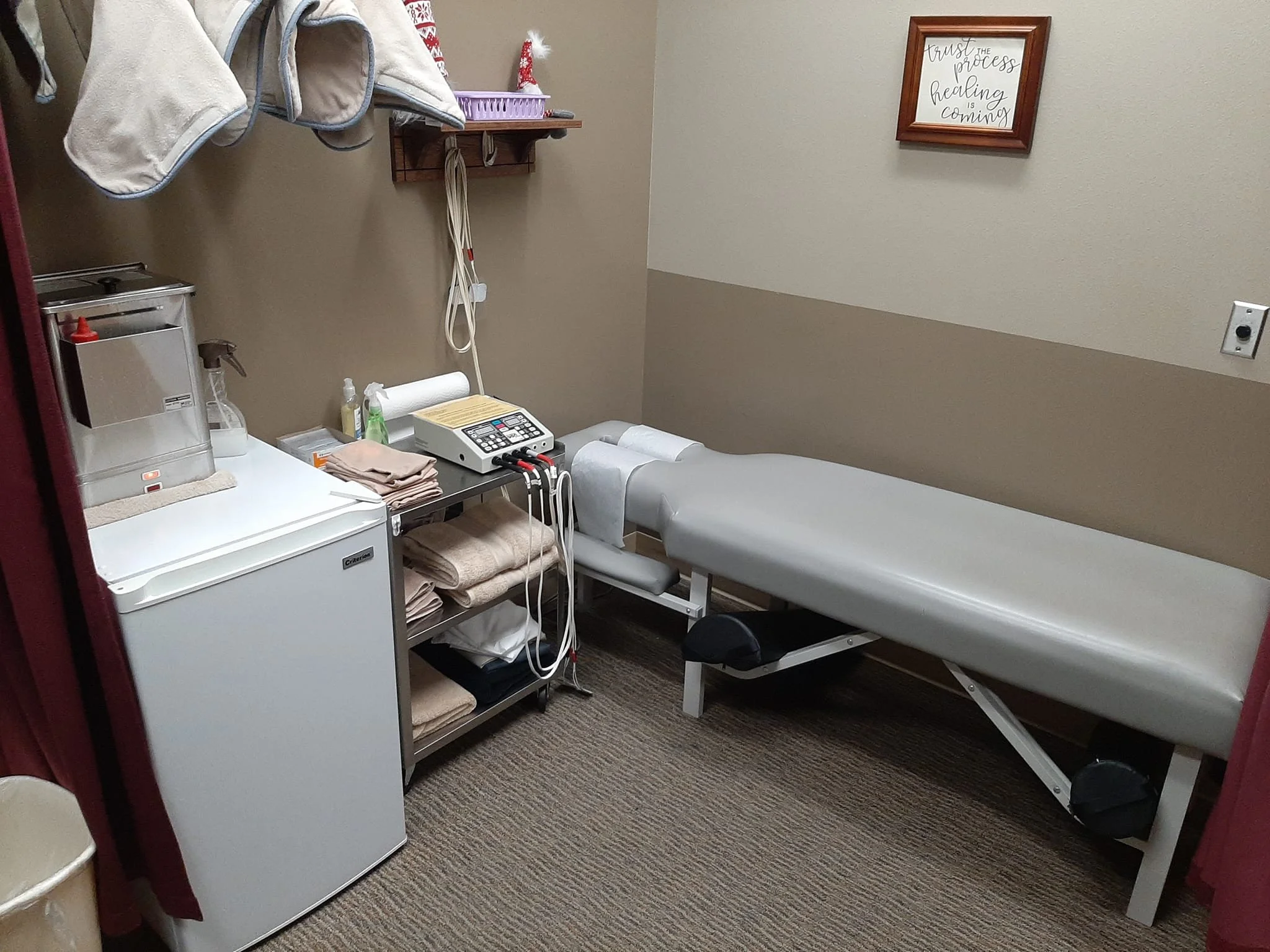 A medical examination room with a gray examination table, a small fridge, a countertop with a tissue box, hand sanitizer, towels, and a medical device with wires. There are shelves with towels and a wall decor sign that reads 'Trust the process, healing is coming'.