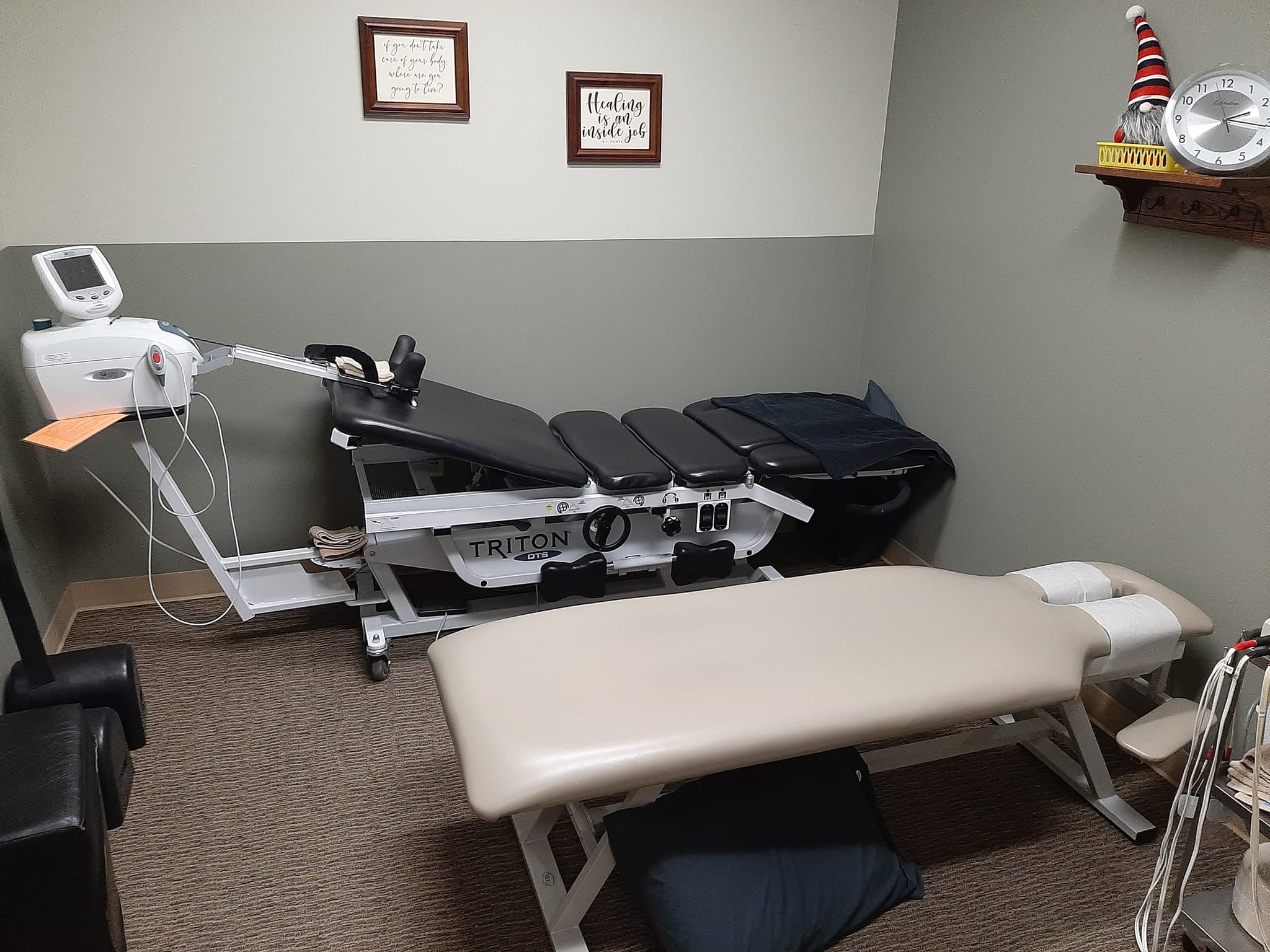Medical examination room with an examination table, a CPT therapy table, medical equipment, wall art, a clock, and holiday decorations