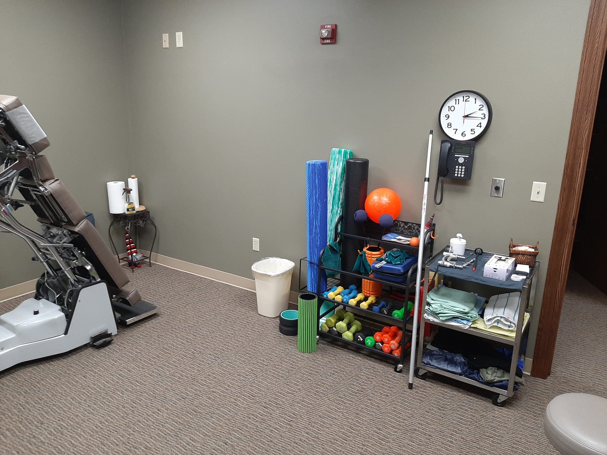Physical therapy or rehabilitation room with exercise equipment, including chair with adjustable parts, white trash bin, shelves with foam rollers, resistance bands, dumbbells, and therapy tools, clock on wall, phone, and medical supplies on cart, and beige upholstered chair.