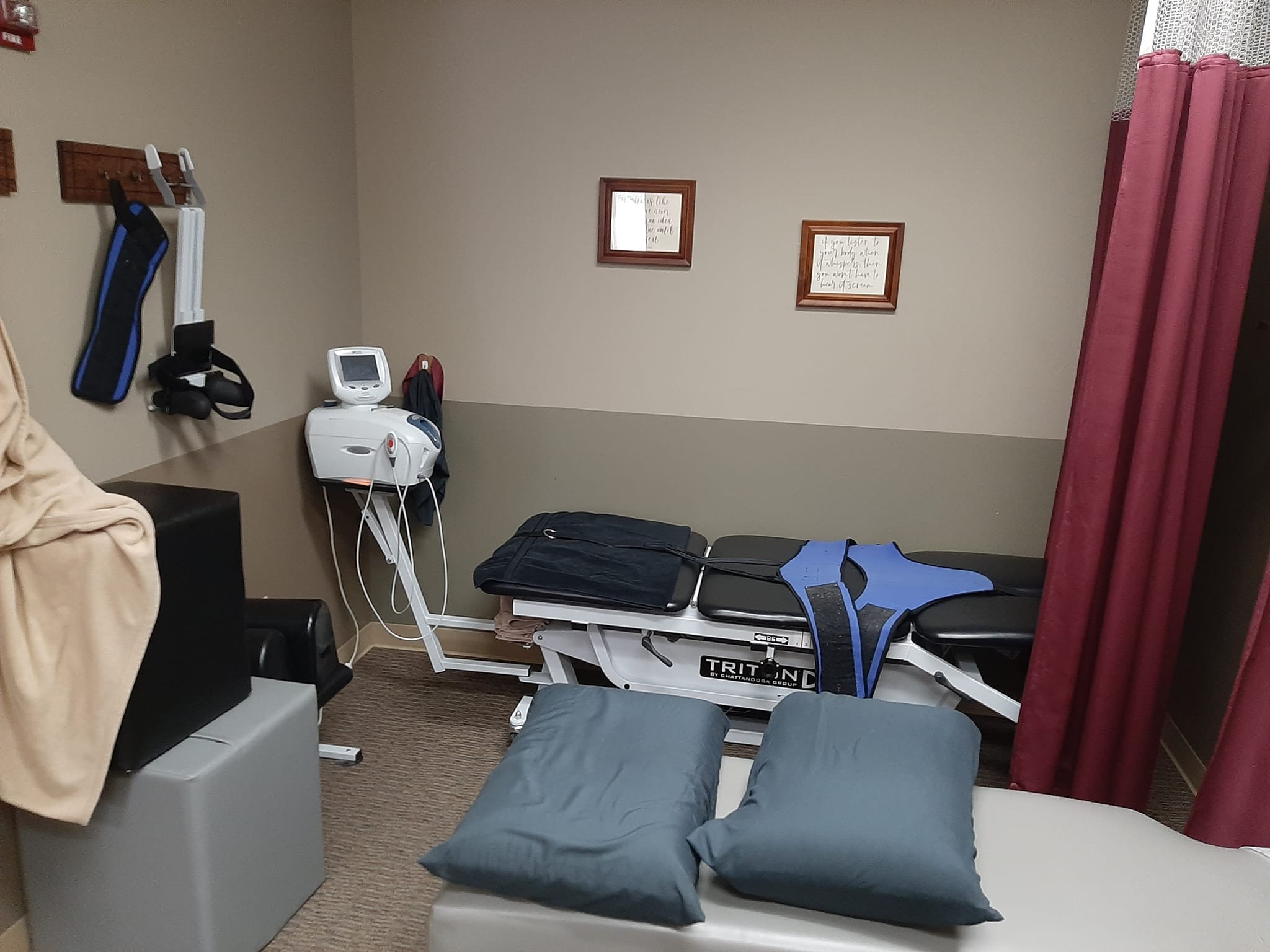A medical examination room with a hospital bed, pillows, medical equipment including a blood pressure cuff and monitor, wall decorations, and privacy curtains.