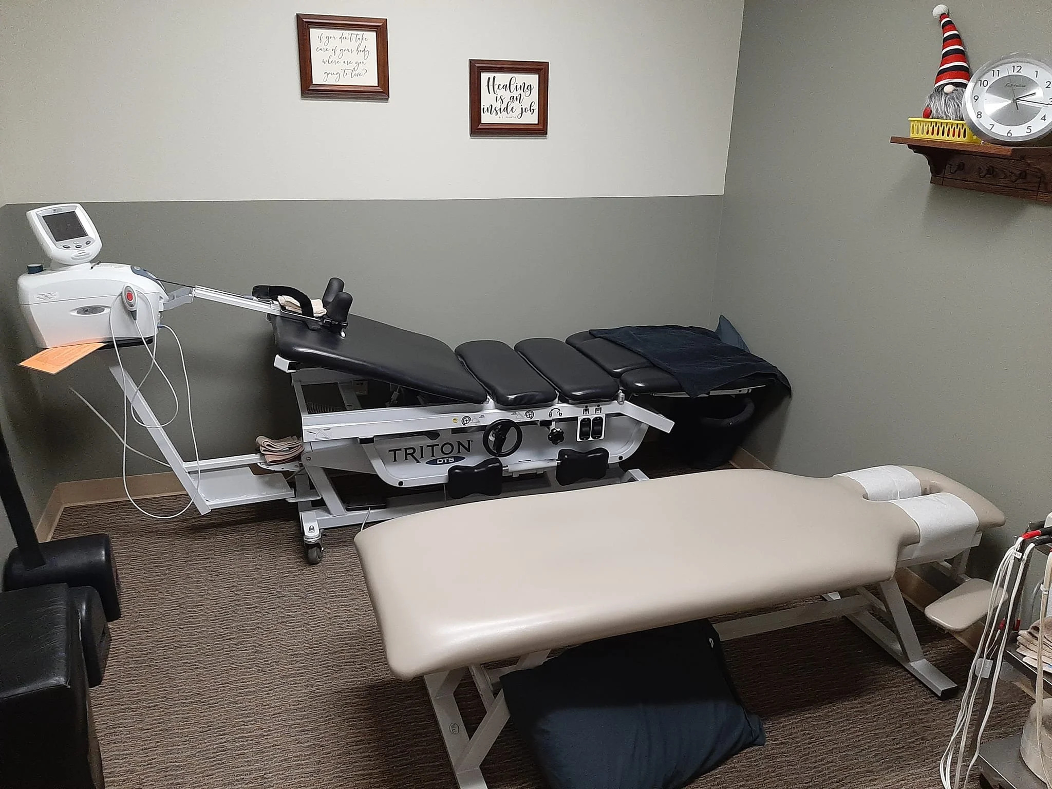 A medical examination room with an examination bed, a therapy table, a wall clock, and wall art. The therapy table has a pillow and black blanket. The wall art includes frames with motivational quotes, and there is a decorative Santa hat and clock on a wall shelf.