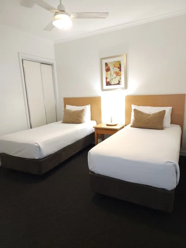 Hotel room with two single beds, each with white sheets and beige pillows, a wooden nightstand with a lamp and phone between the beds, a framed abstract art piece above the nightstand, a ceiling fan, and a closet with sliding doors.