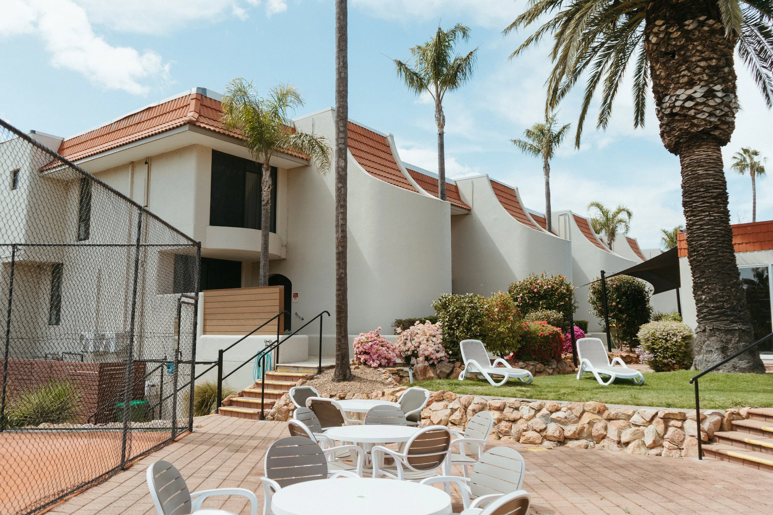 Lake Mulwala Yarrawonga - Exterior view of a white residential complex with curved roofs, surrounded by palm trees and landscaped garden with pink and white flowers, outdoor tables and chairs, and lounge chairs.