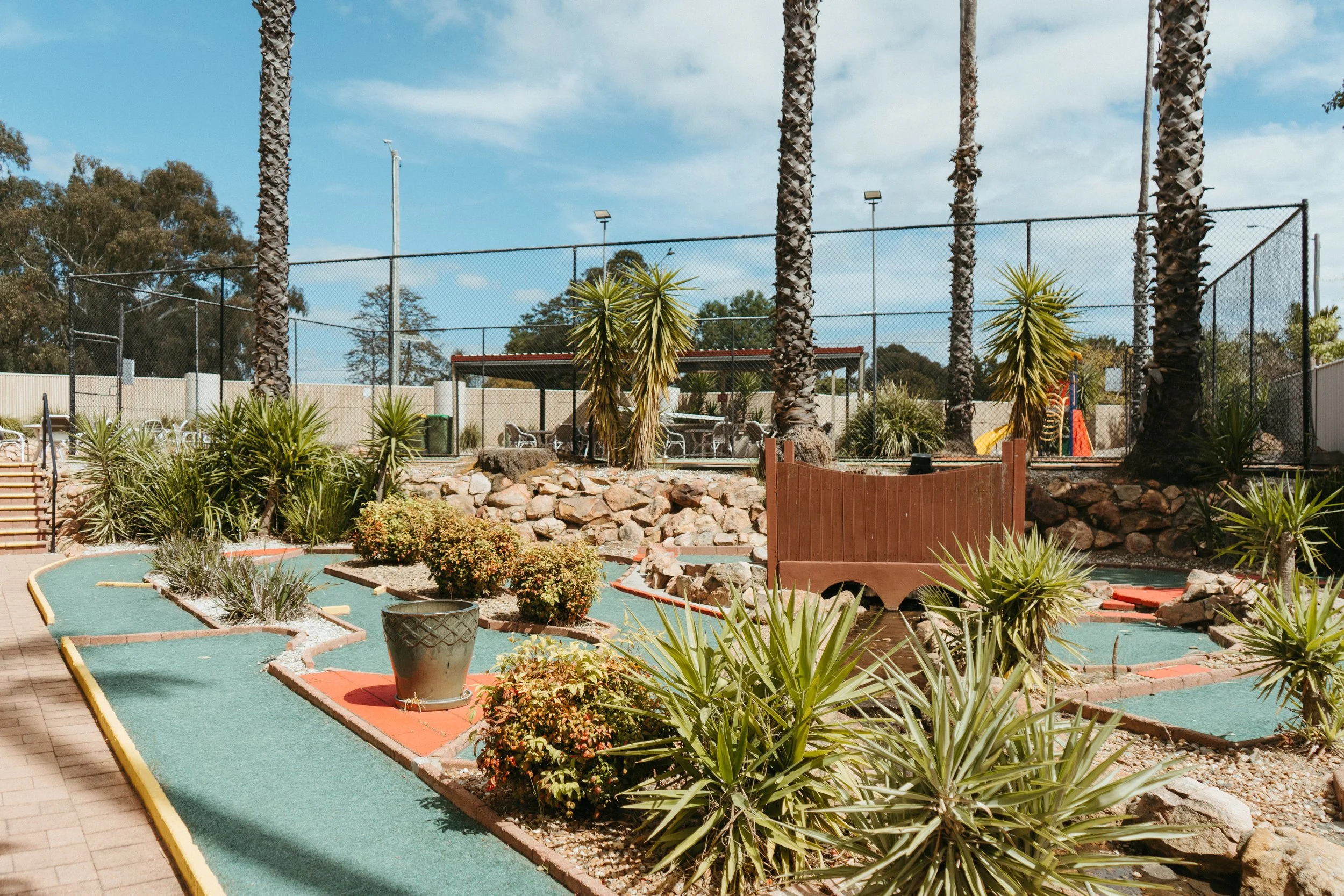 Lake Mulwala Yarrawonga - Miniature golf course with palm trees, plants, and a fenced, sunny outdoor area with a playground in the background