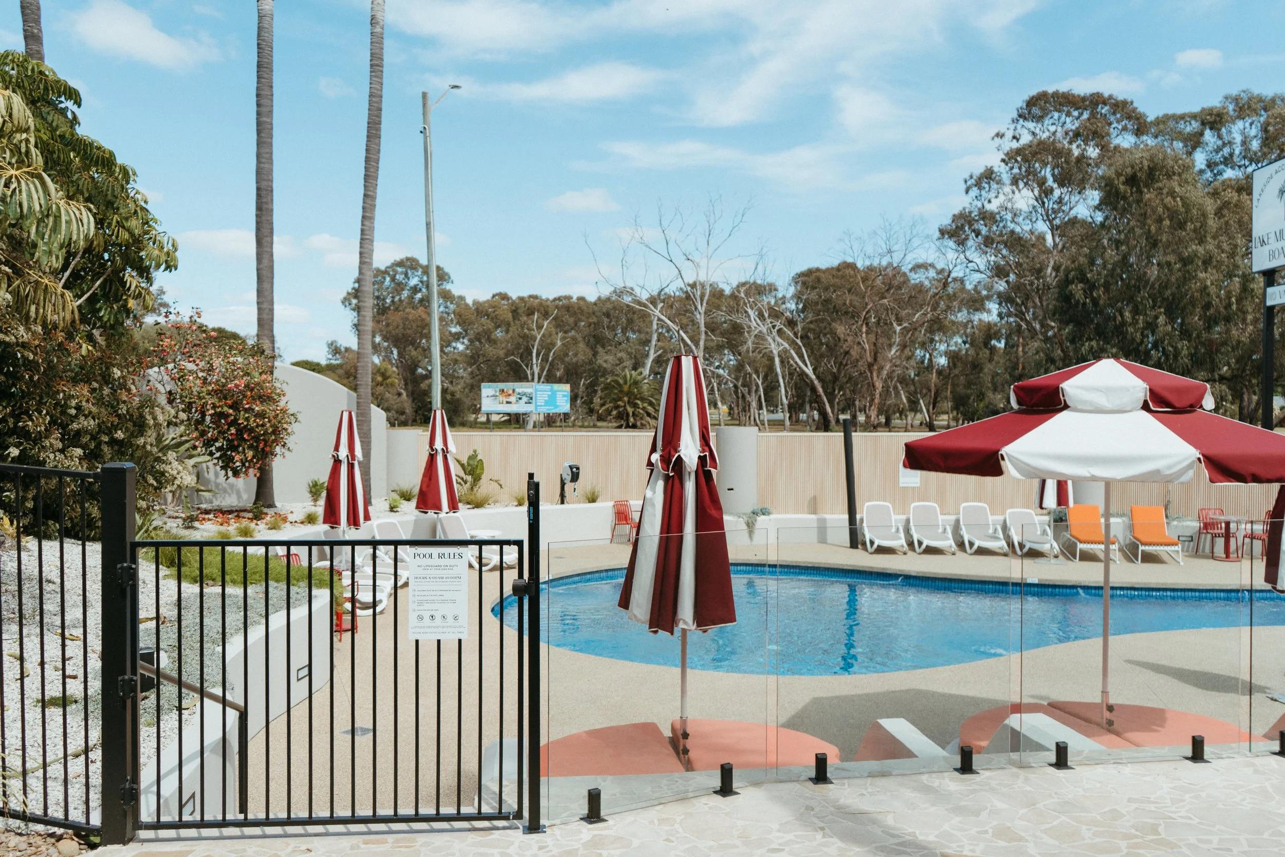 Lake Mulwala Yarrawonga - outdoor swimming pool area with closed red and white striped umbrellas, lounge chairs, and a glass barrier with a sign. Surrounded by trees and plants, sunny weather with partly cloudy sky.