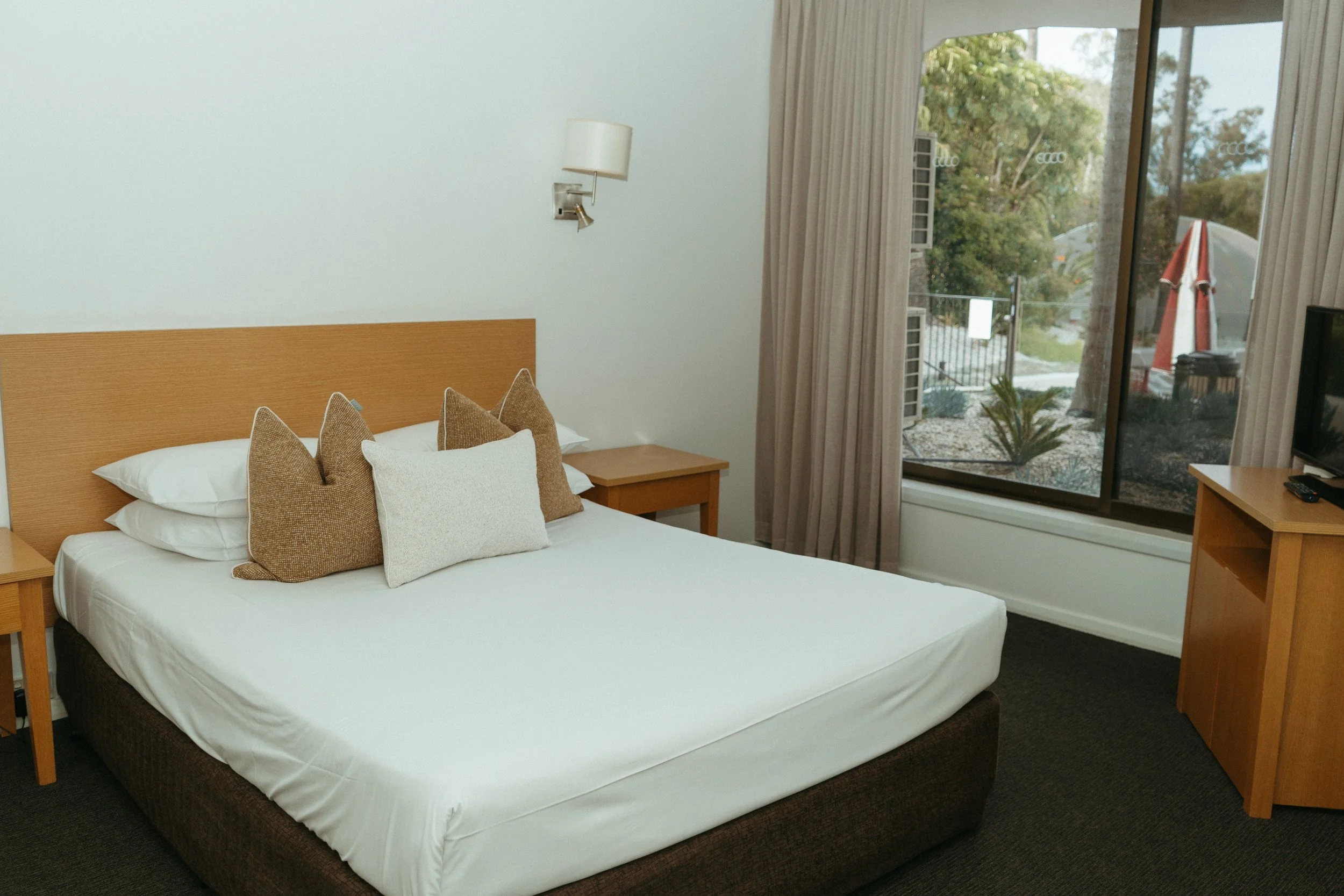 Lake Mulwala Yarrawonga - Hotel room with a bed, bedside tables, a wall-mounted lamp, a window with curtains, a television on a wooden stand outside the window, and outdoor scenery including trees and an outdoor umbrella.
