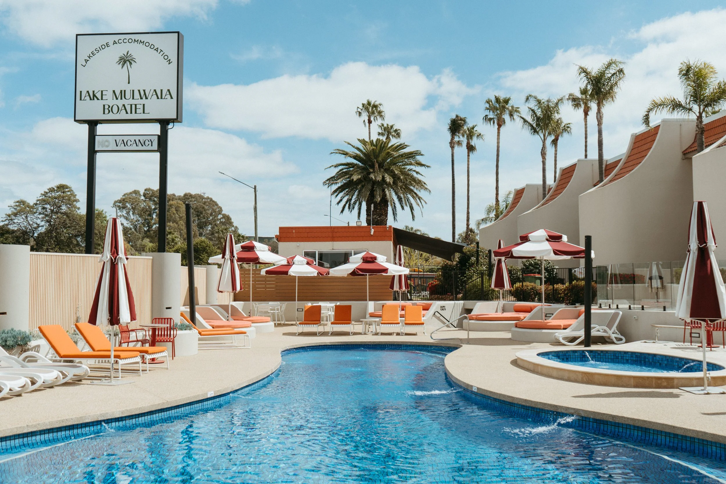 Poolside area with orange lounge chairs, white tables, red and white umbrellas, and a small round hot tub. There is a sign reading 'Lake Mulwala Boatel' with a palm tree logo. Tall palm trees and buildings are in the background under a partly cloudy 