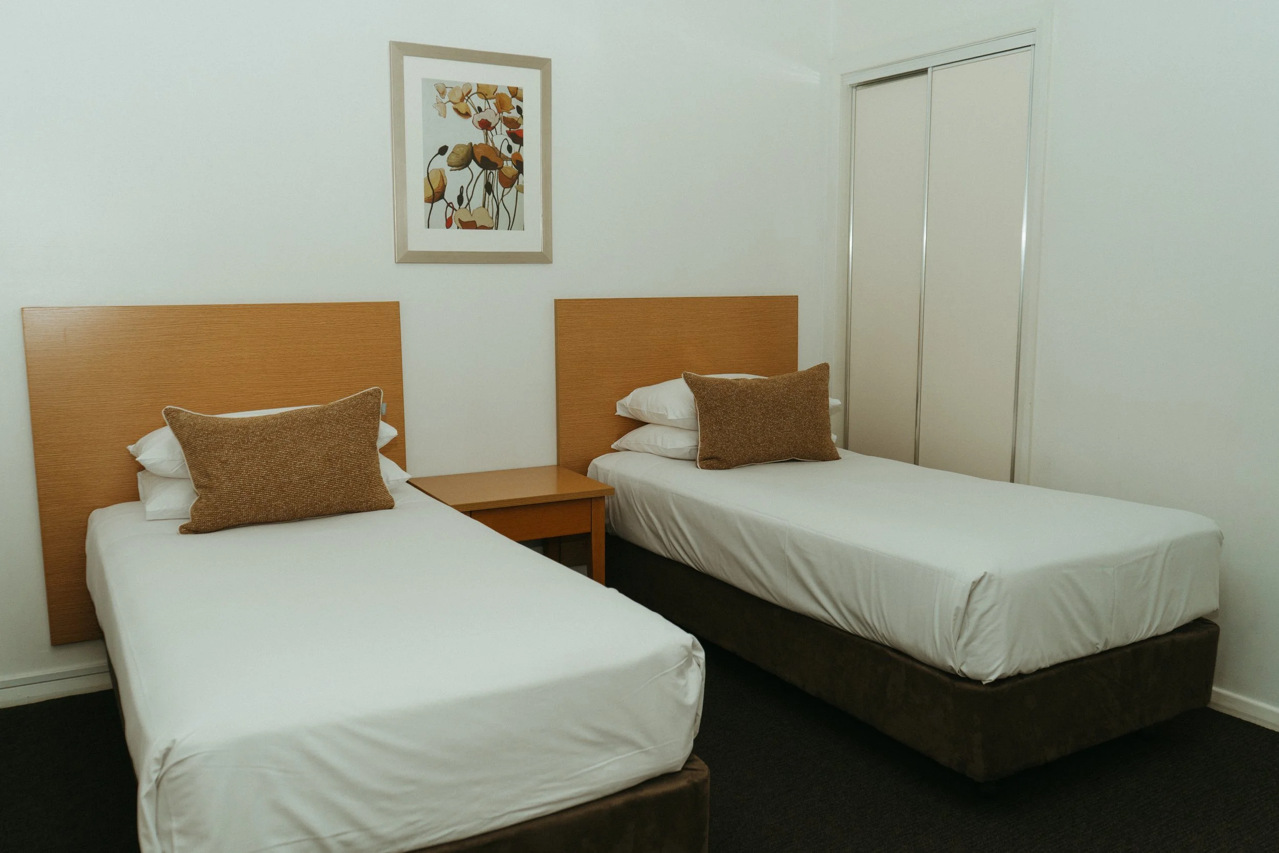 Lake Mulwala Yarrawonga - Hotel room with two twin beds, each with a brown pillow, a wooden headboard, a nightstand, a framed floral artwork above the beds, and a closet with sliding doors.