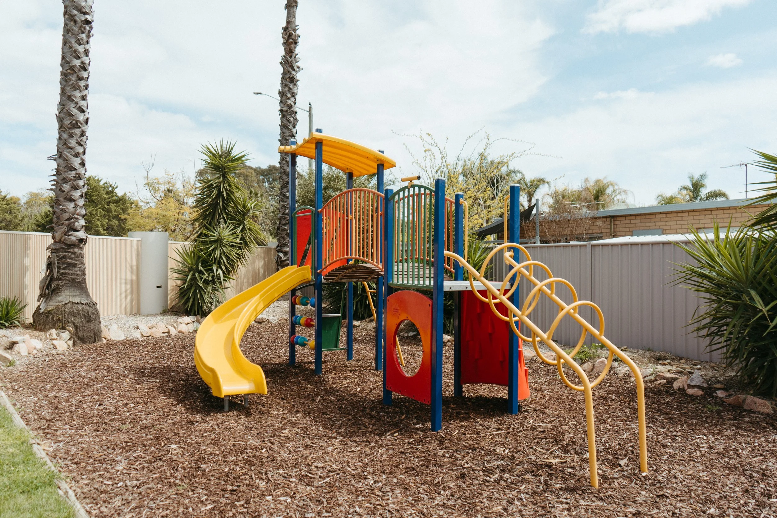 Lake Mulwala Yarrawonga - Colorful outdoor playground with slides, climbing structures, and monkey bars, surrounded by palm trees and a fenced yard.