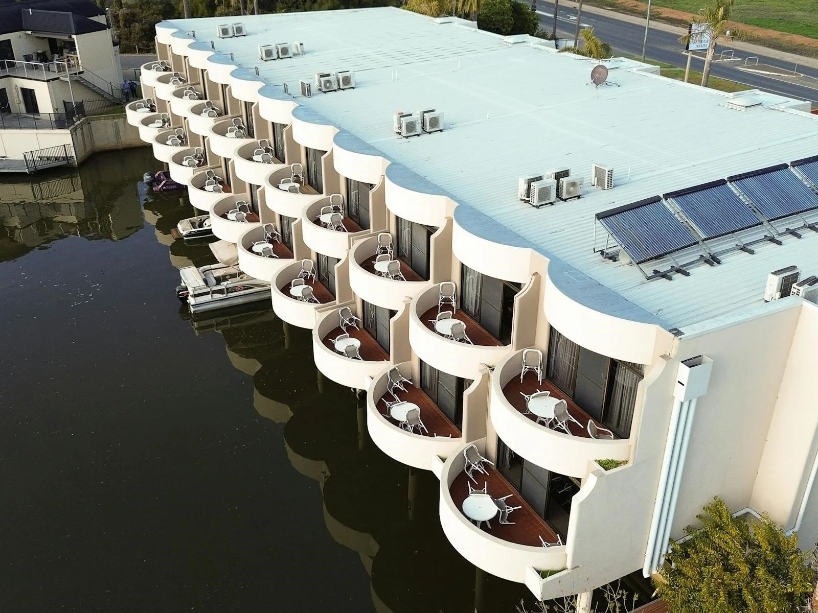 Lake Mulwala Yarrawonga - Multiple small balconies with white chairs and tables extend over a waterway from a modern building with a flat, light-colored roof, solar panels, and air conditioning units.