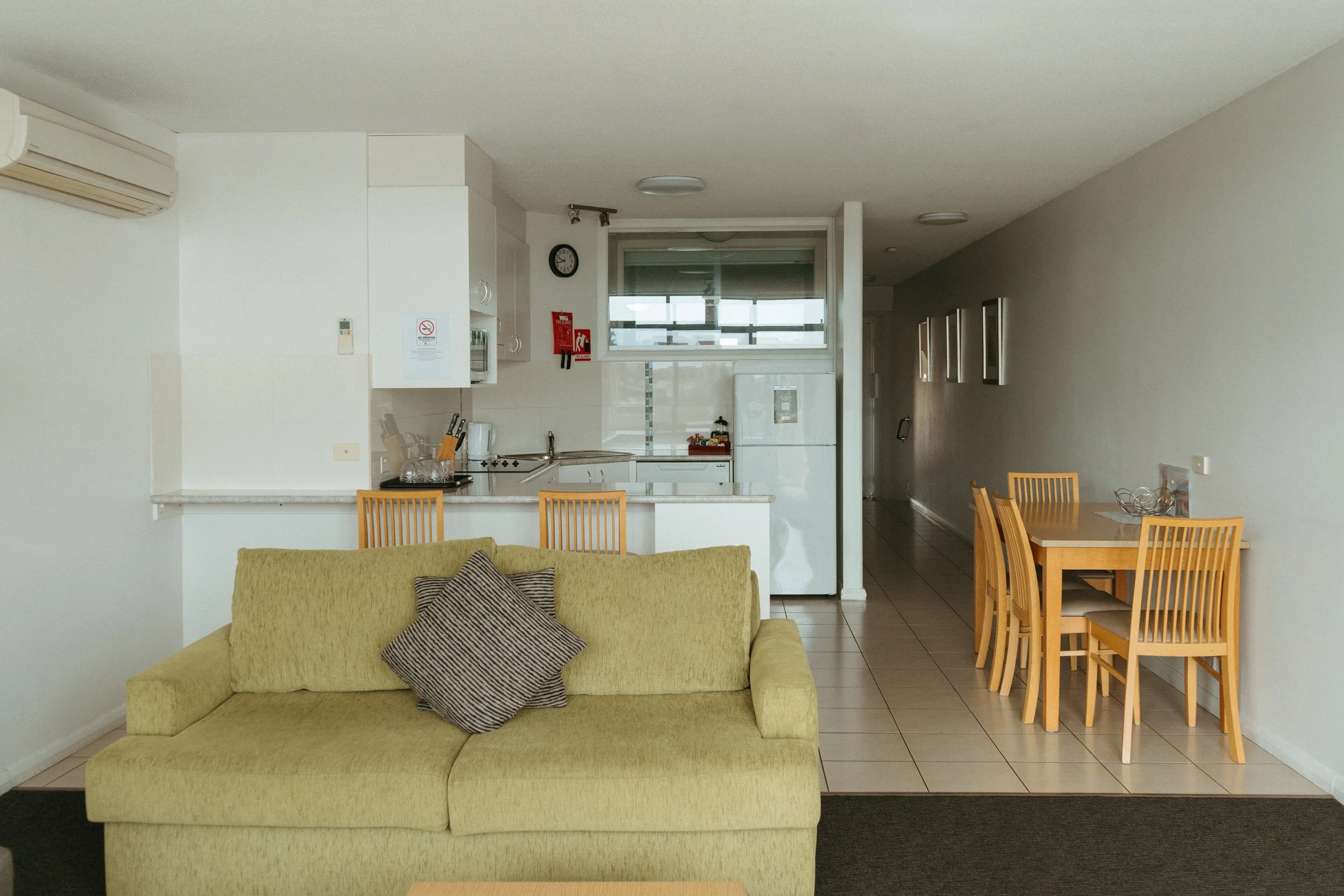 Lake Mulwala Yarrawonga - Living room with a yellow sofa and a pillow, a dining table with six chairs, a kitchen with appliances, a window, and hallway.
