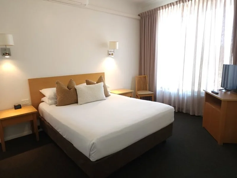A hotel room with a bed, pillows, two nightstands, a chair, and a television near a large window with curtains.