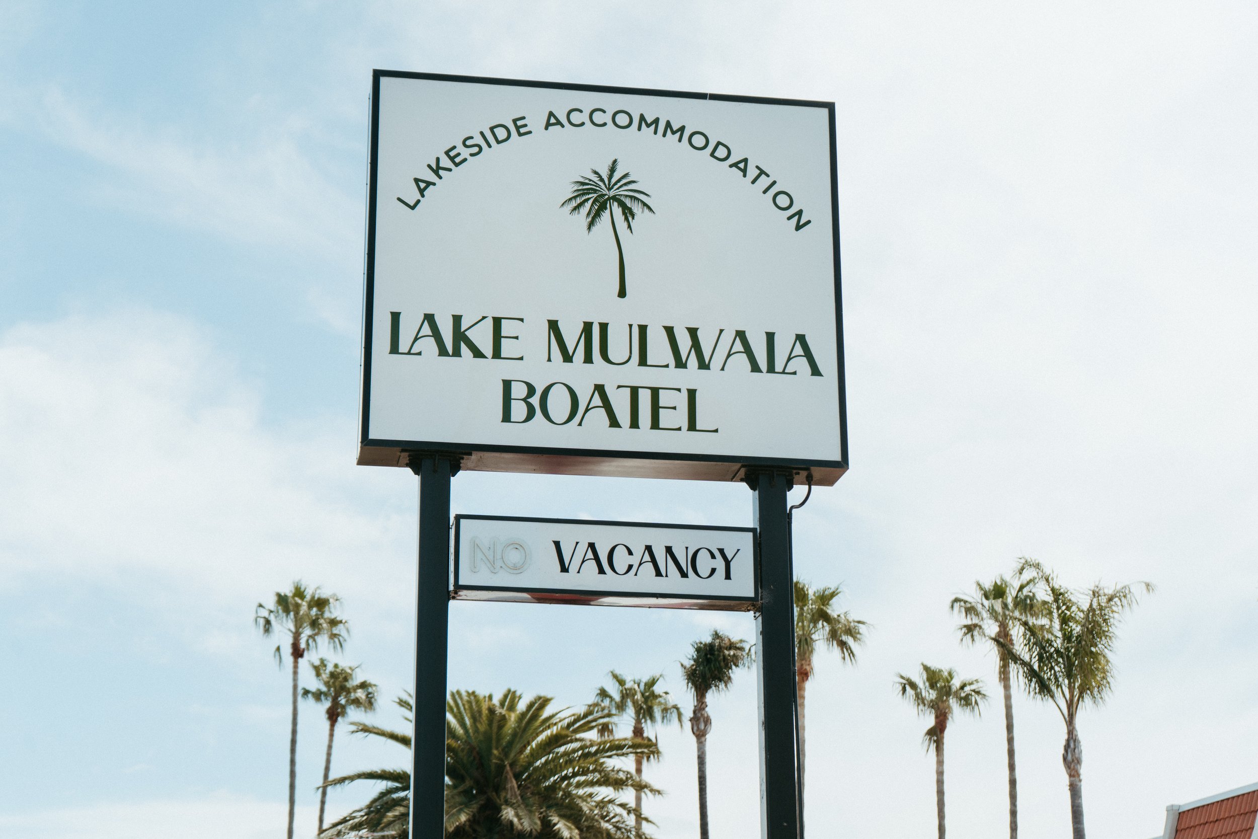 Lake Mulwala Yarrawonga - Sign for Lakeside Accommodation Lake Mulwala Bateau with a palm tree logo, and a smaller sign below reading 'No Vacancy', in front of palm trees and a cloudy sky.