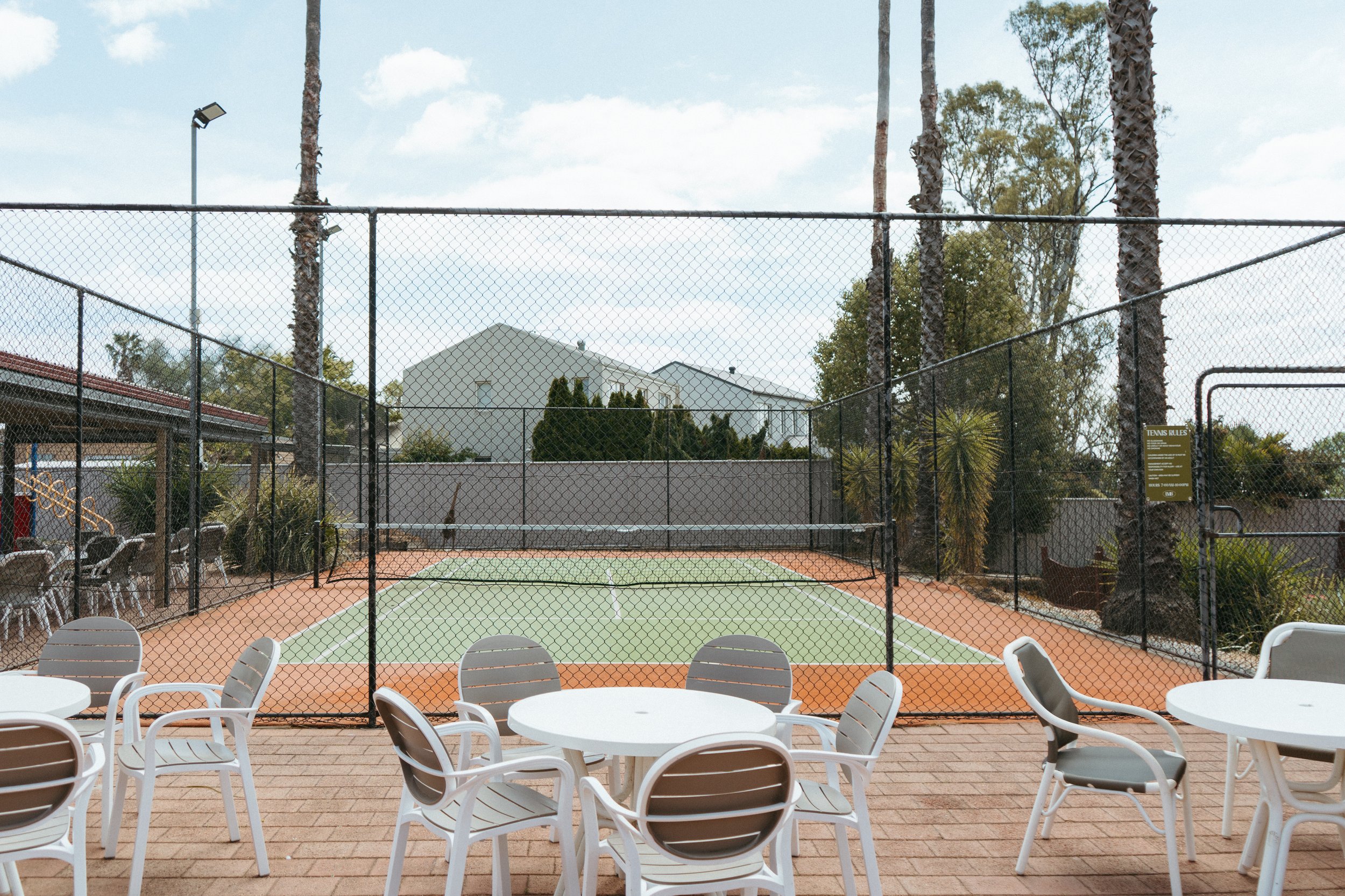 Lake Mulwala Yarrawonga - Outdoor tennis court surrounded by a black chain-link fence with patio tables and chairs in the foreground.