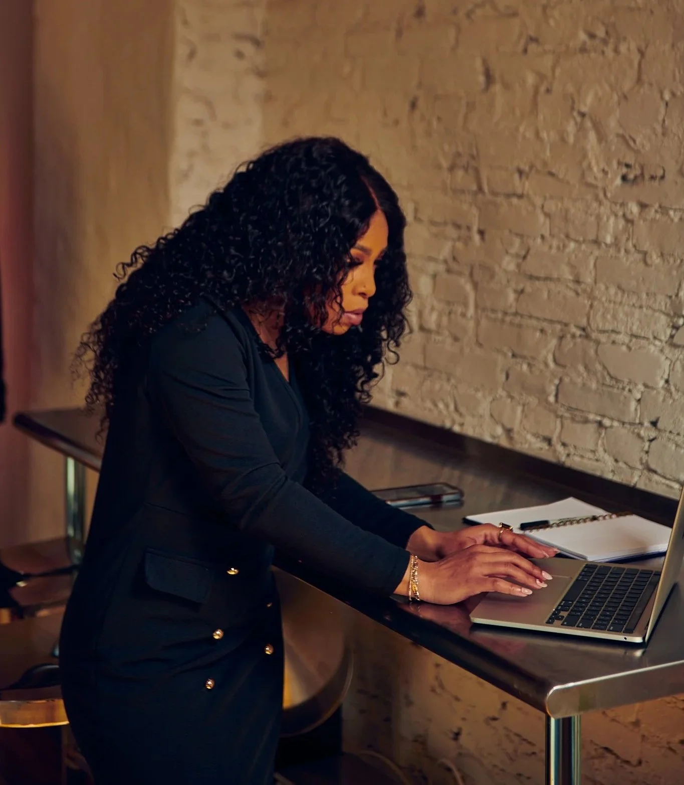 A woman with curly black hair working on a laptop at a bar-height table in a brick-walled room.