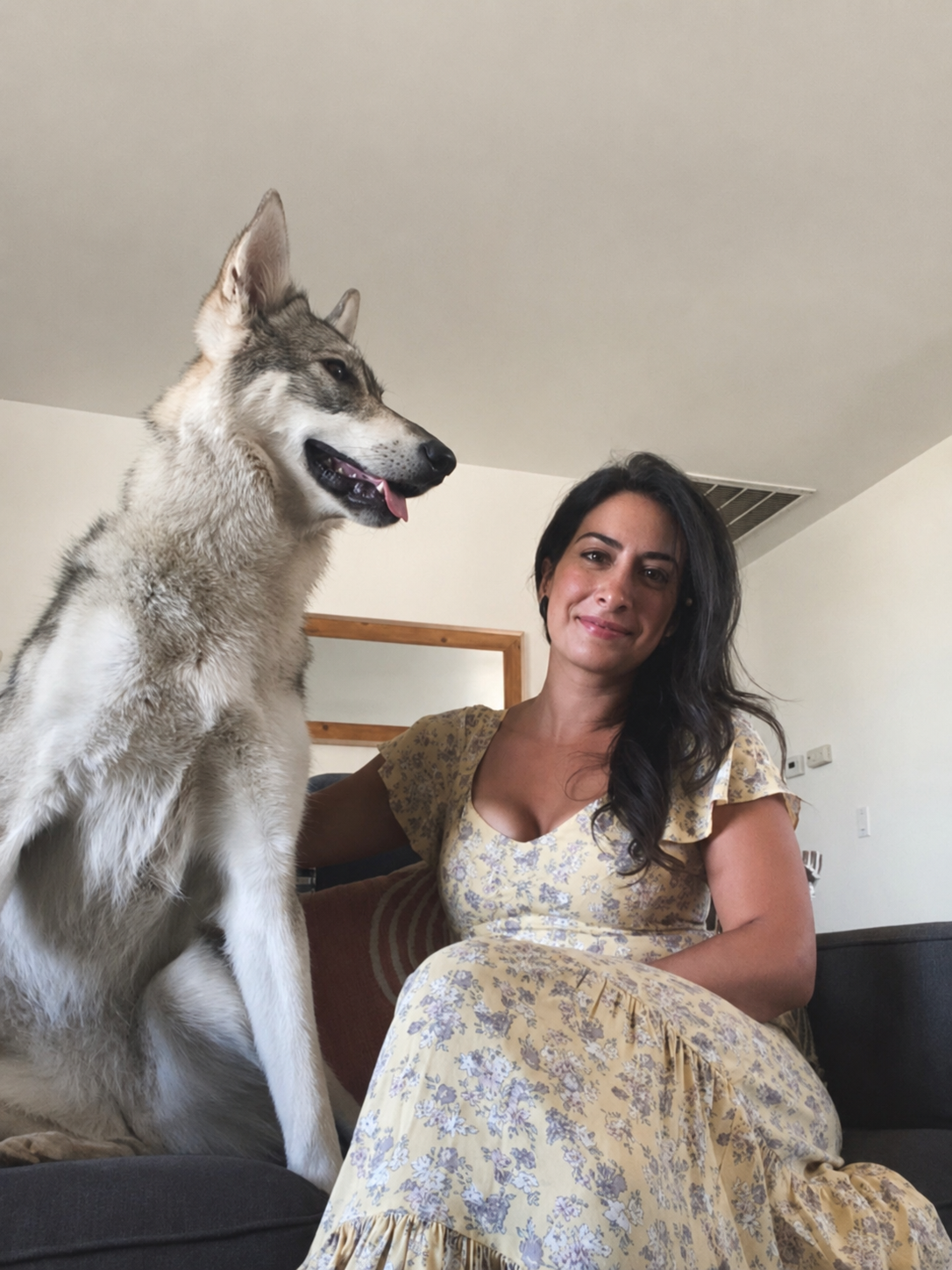 A woman with long dark hair sitting on a couch next to a large Siberian Husky dog.