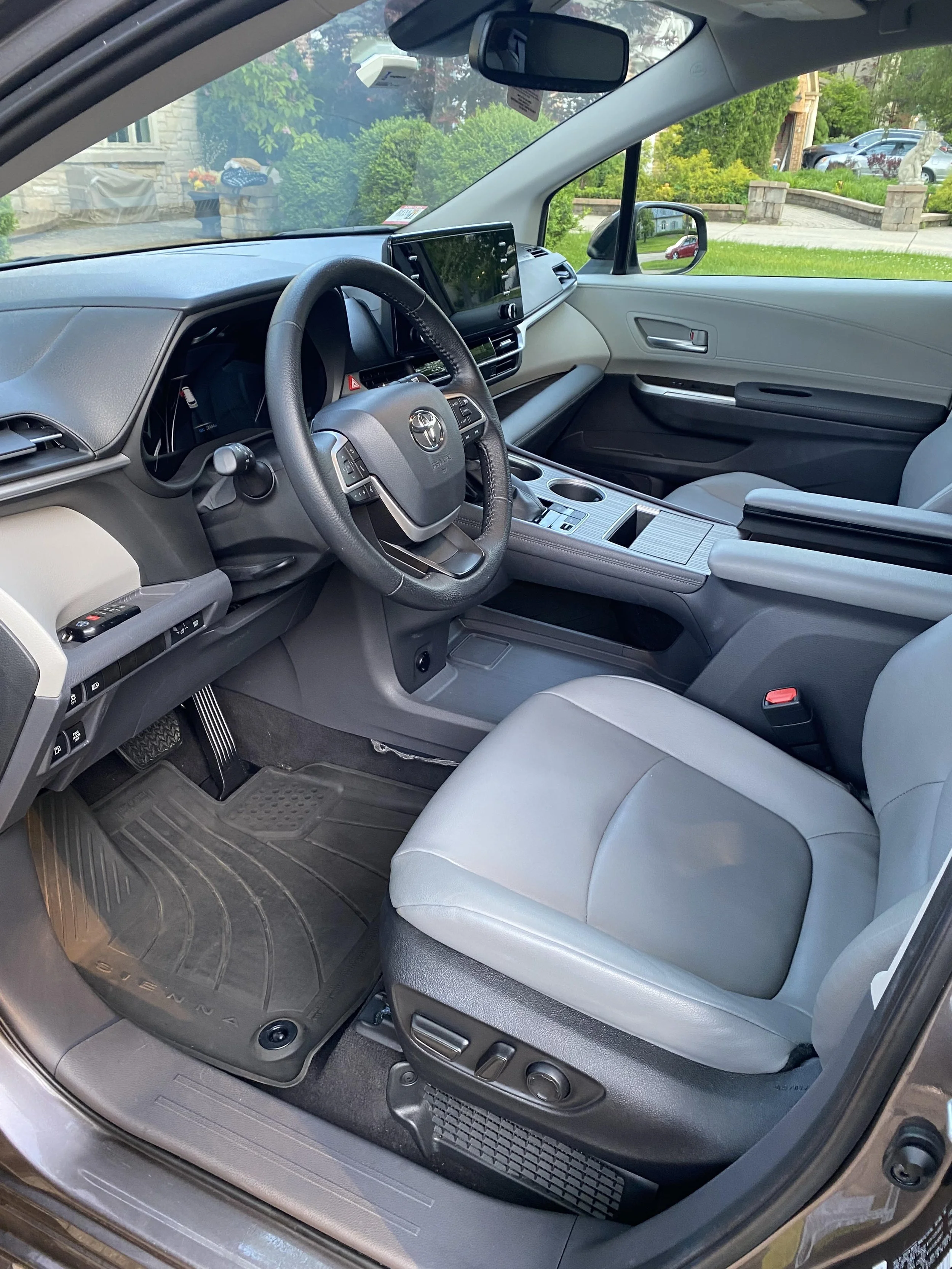 Interior of a modern car showing the driver's seat, steering wheel, dashboard, and center console with a touchscreen display. The car features gray leather seats and a black floor mat labeled 'Sienna.'