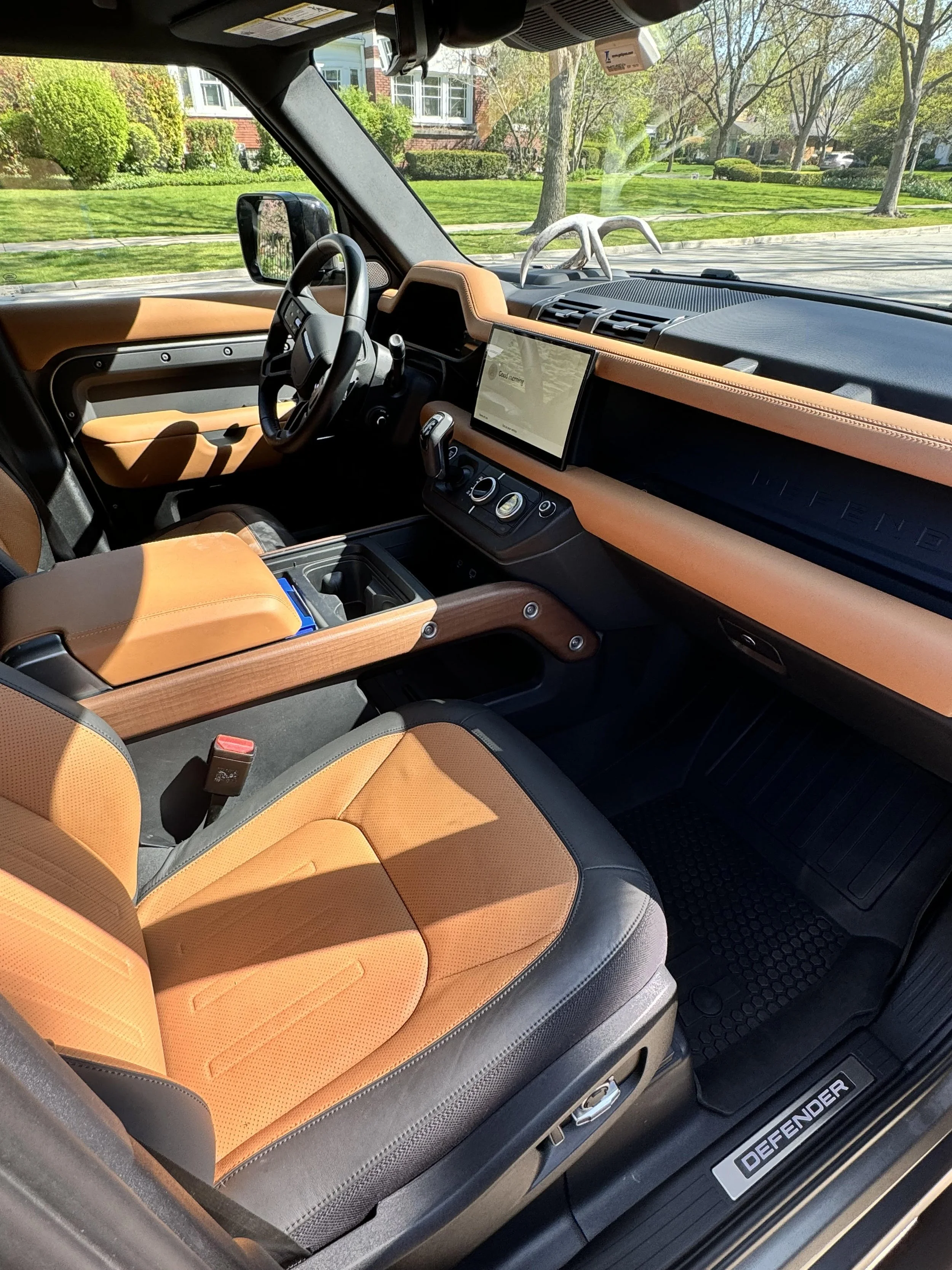 Interior of a car with tan leather seats and dashboard, black steering wheel, digital display, and controls. Sunlight is streaming through the window, highlighting the modern design.