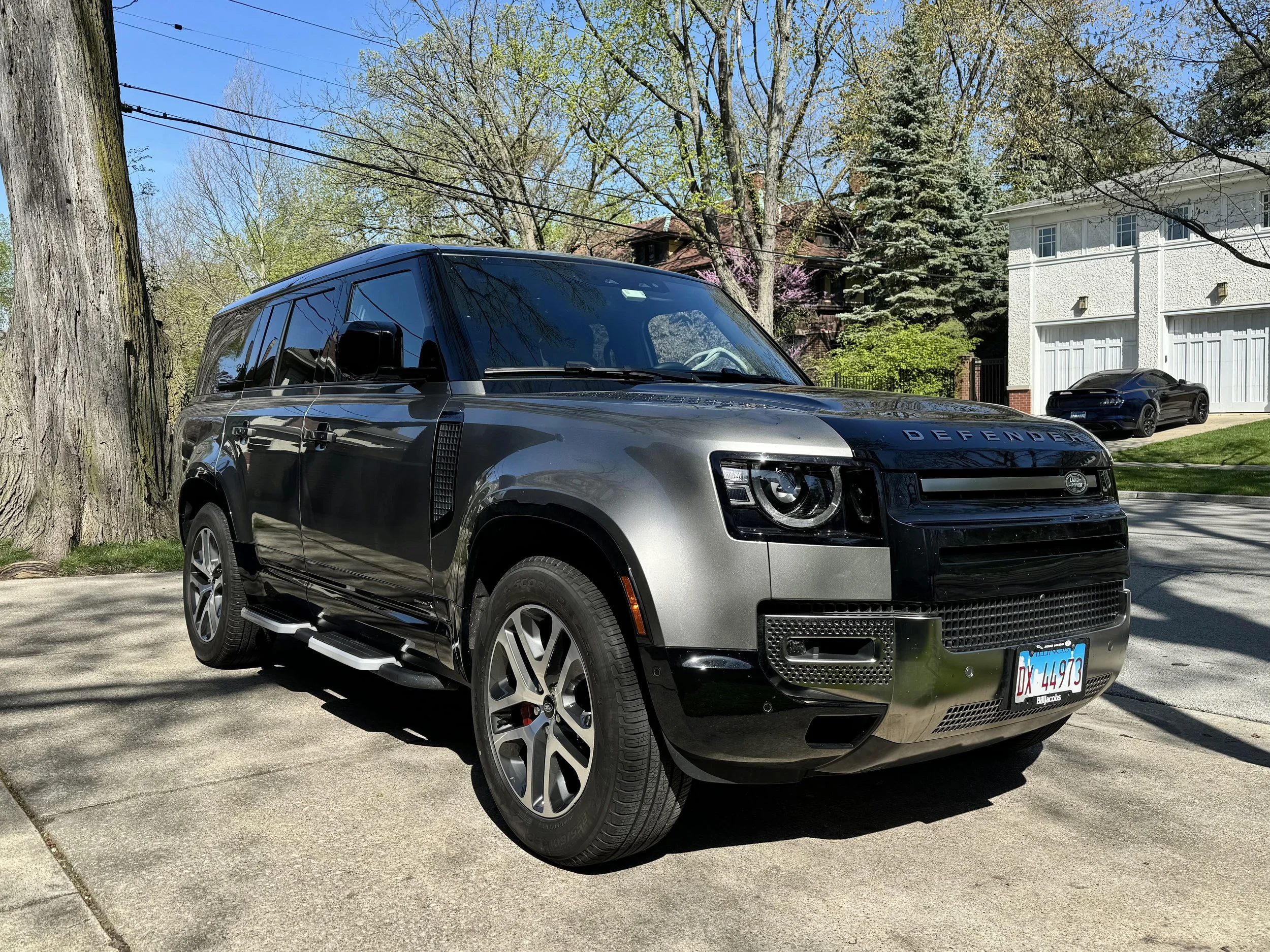 Gray Land Rover Defender SUV parked on a driveway in a suburban neighborhood with trees and houses in the background.