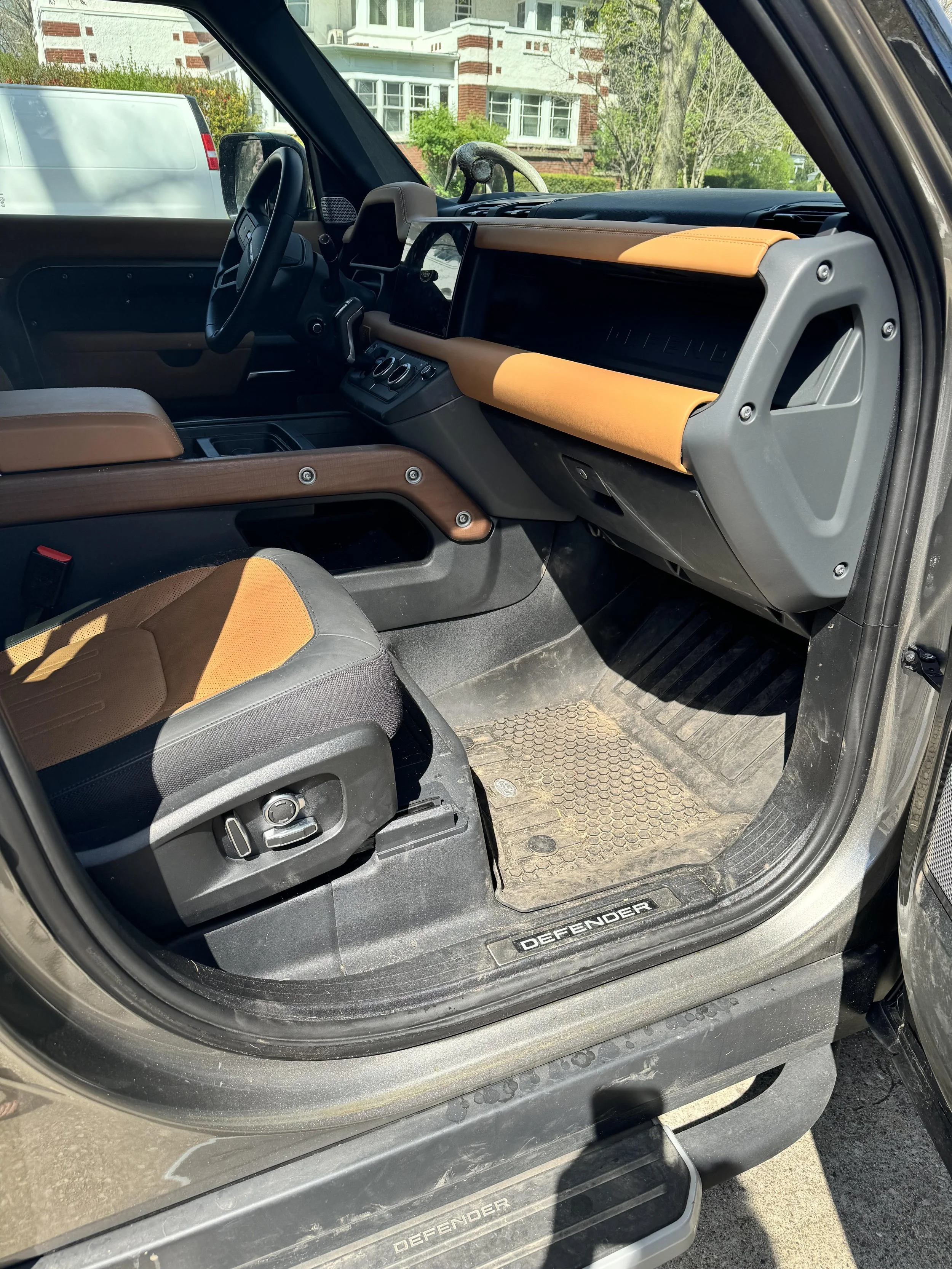 Interior of a Land Rover Defender showing the driver's seat, dashboard, and steering wheel with tan and black upholstery in a parked vehicle.