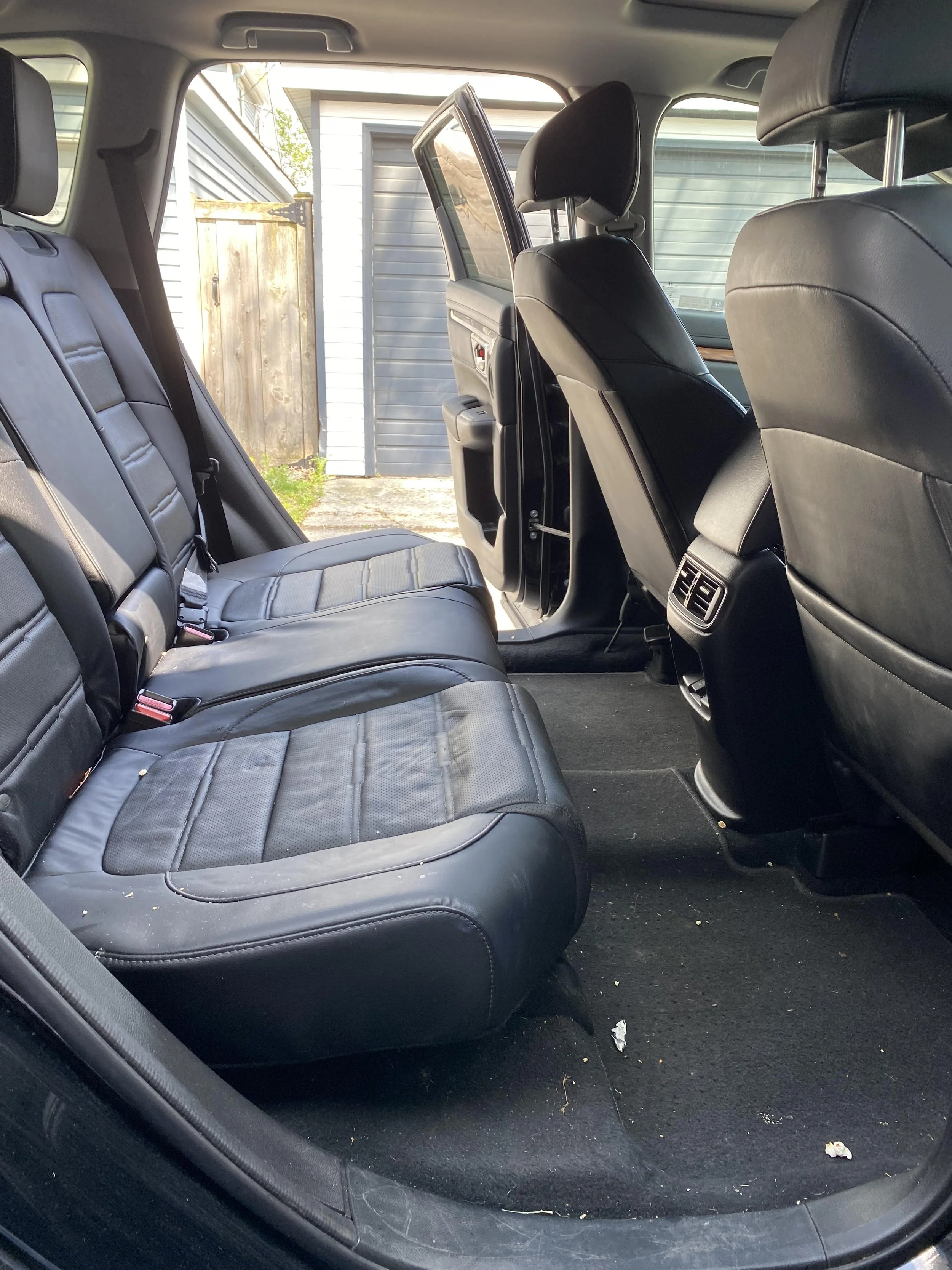Interior of a car with black leather seats, open rear door, and visible garage and wooden fence outside.