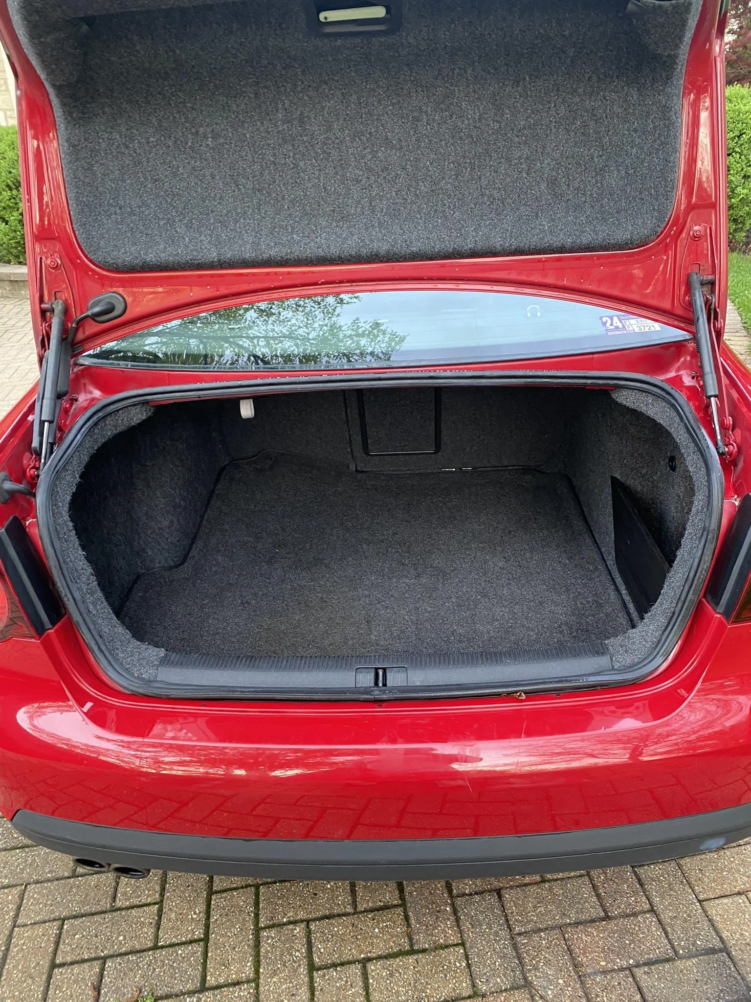 Open trunk of a red car, showing an empty carpeted interior, parked on a brick driveway.