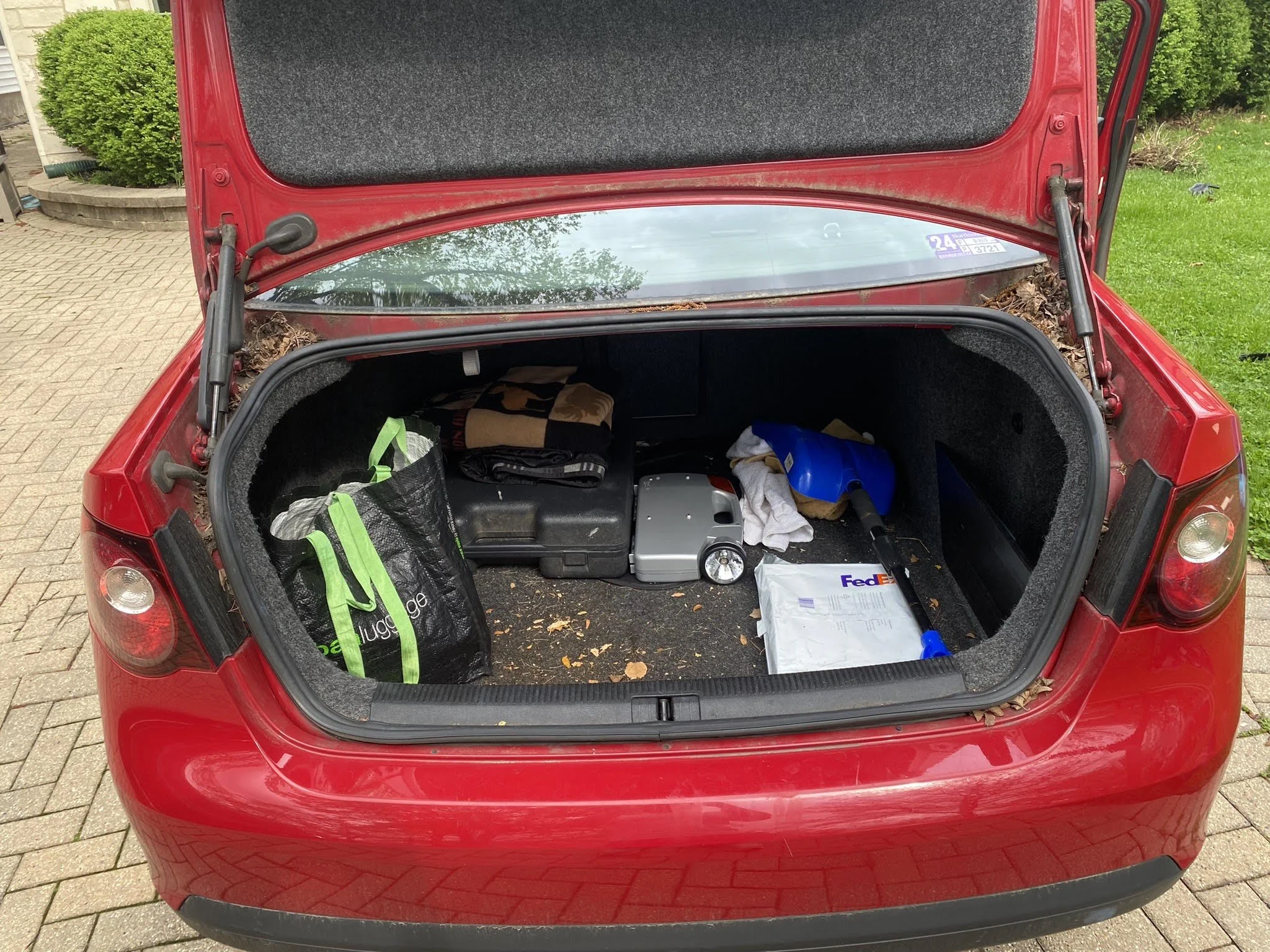 Open trunk of a red car containing a green reusable shopping bag, leaf debris, a blue shovel, a suitcase, a FedEx envelope, and a brown box.