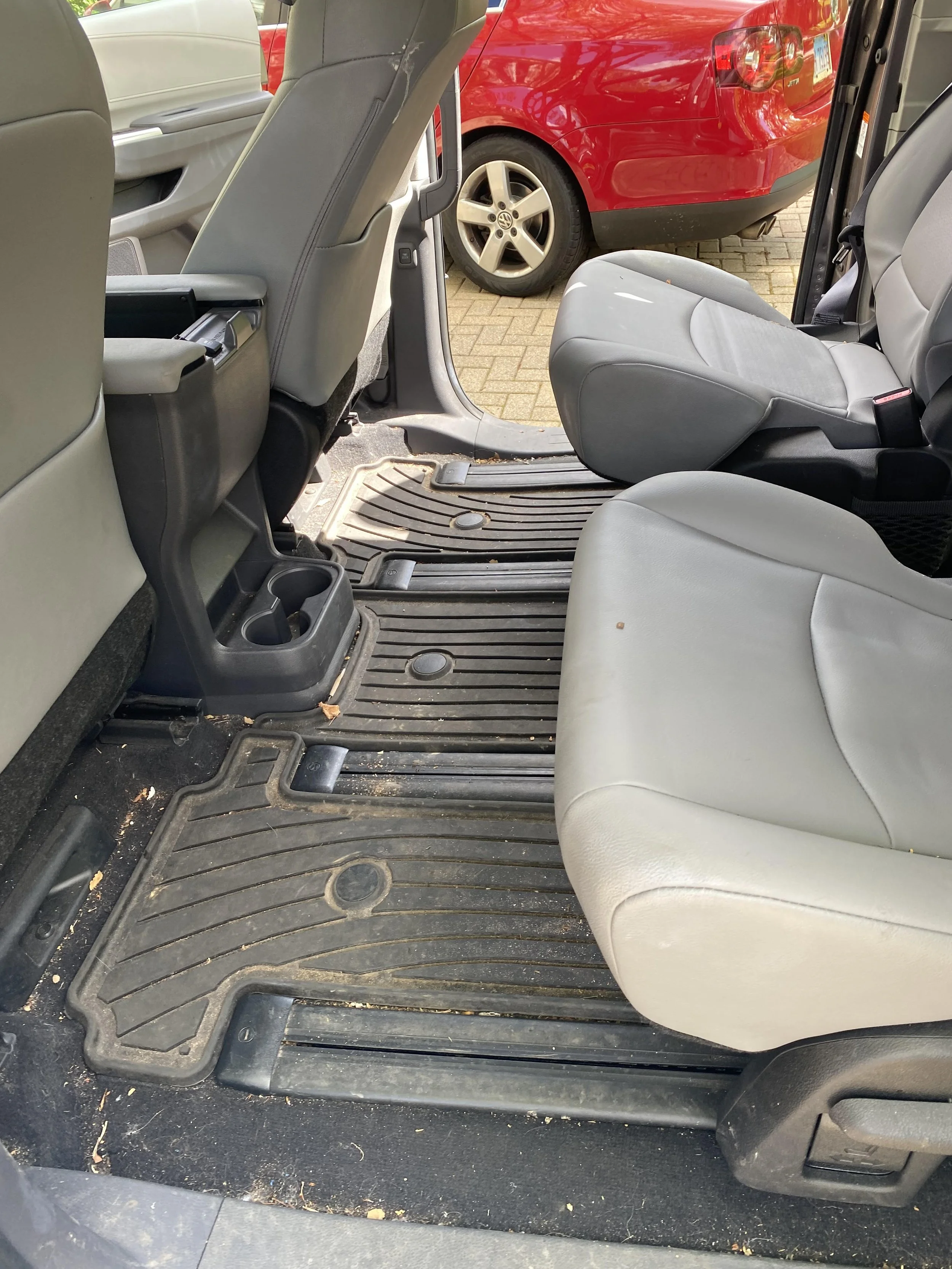 Interior of a car showing the rear and front passenger seats with rubber floor mats and a cup holder, visible door panel and part of a red car outside.