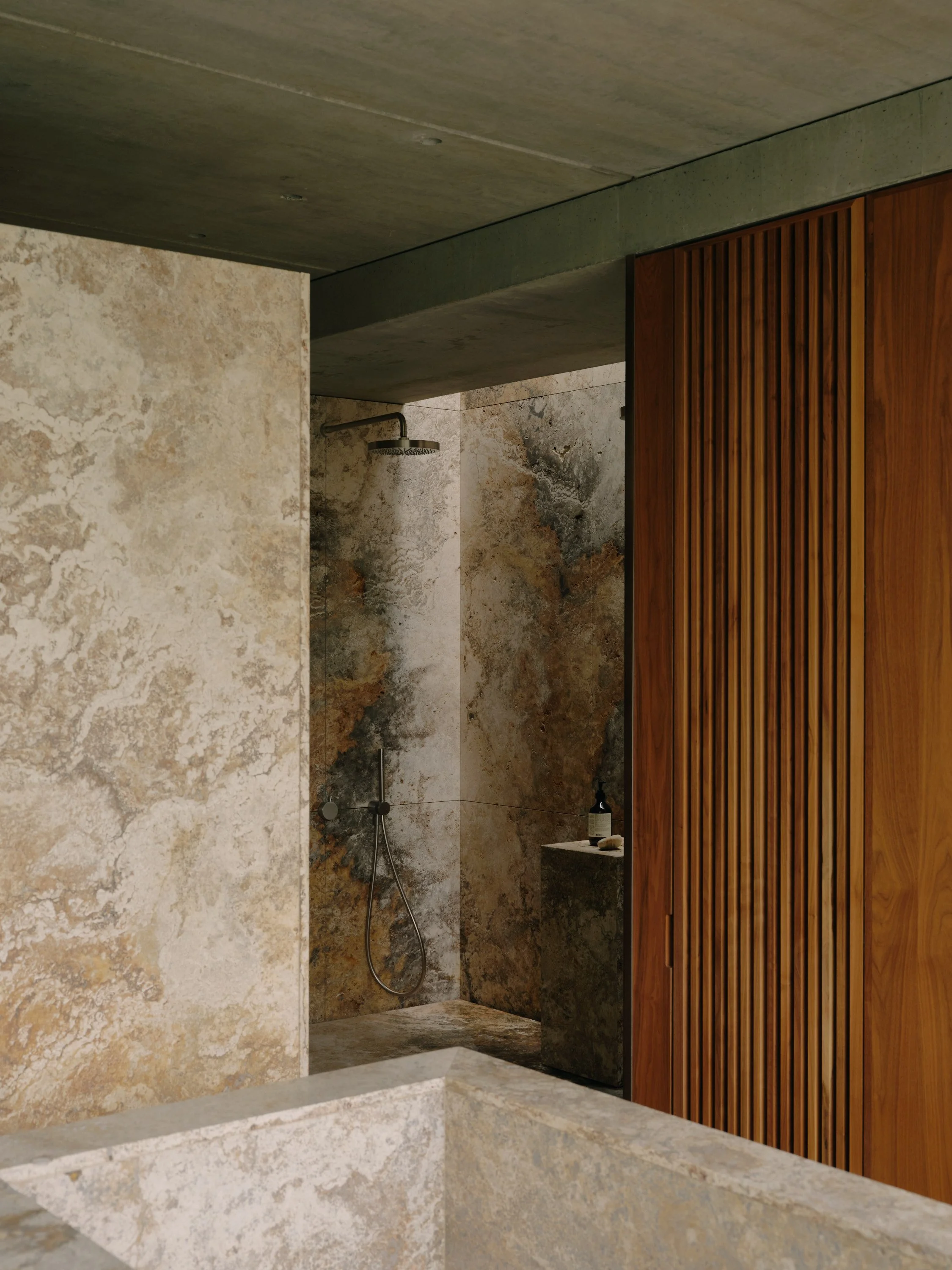 Interior view of a modern bathroom featuring a walk-in shower with Travertine slab walls, a rainfall shower head, and Walnut louvered privacy panel.