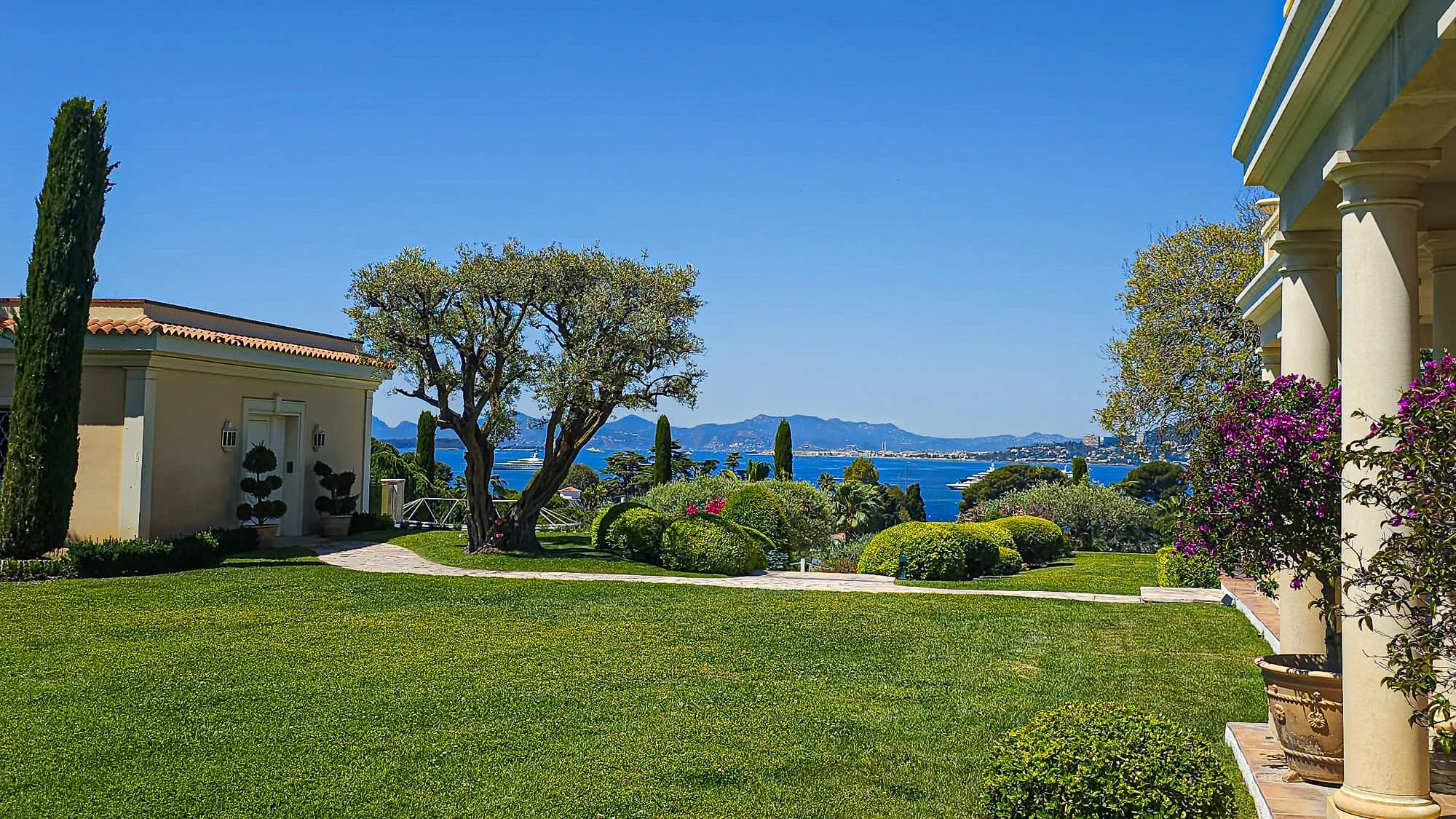 Jardin bien entretenu avec pelouse verte, arbres et hortensias en fleurs, vue sur la mer et montagnes au loin, côté d'une grande maison avec colonnes et volets bleus.