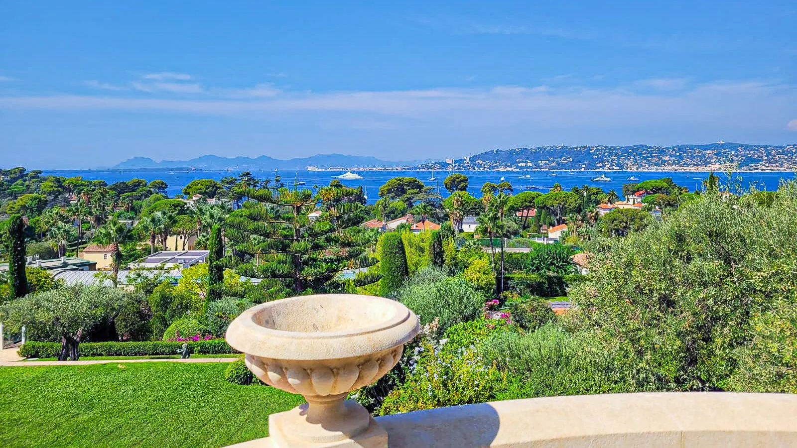 Vue panoramique d'une côte méditerranéenne avec mer bleue, yachts, collines, et végétation luxuriante, vue depuis un balcon avec un soubassement en pierre.