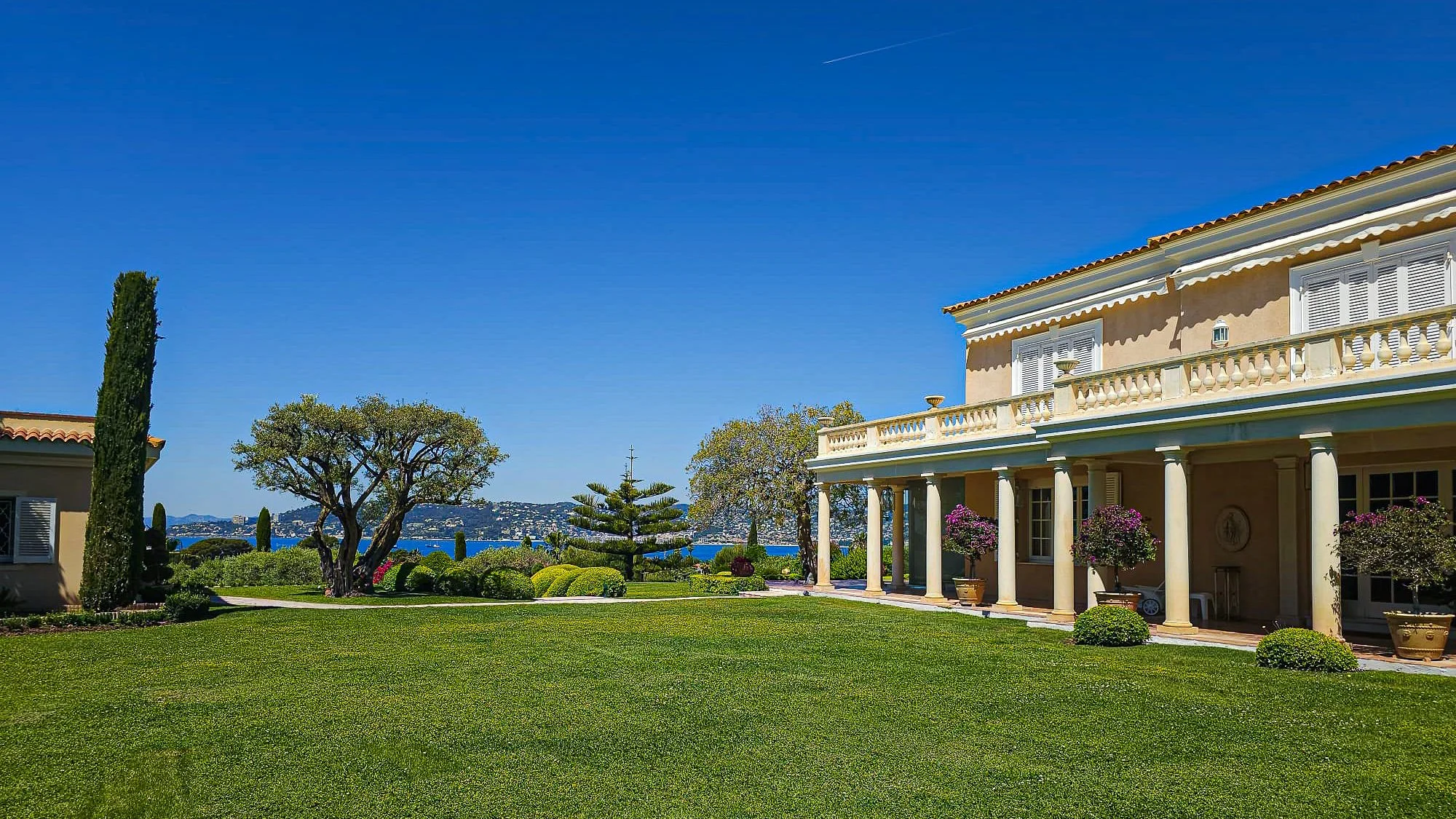 Grande maison style villa avec colonnes et balustrades cachée derrière un jardin bien entretenu, arbres et haies, avec vue sur la mer au fond sous un ciel bleu clair.