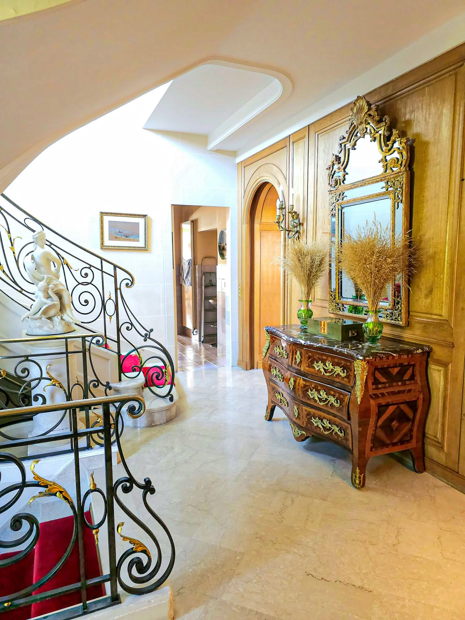 Intérieur d'une maison avec escalier en fer forgé, statue en marbre, tableau dans un cadre doré, meuble ancien en bois avec des décorations en or, miroir avec cadre ornemental, plantes en vases verts.