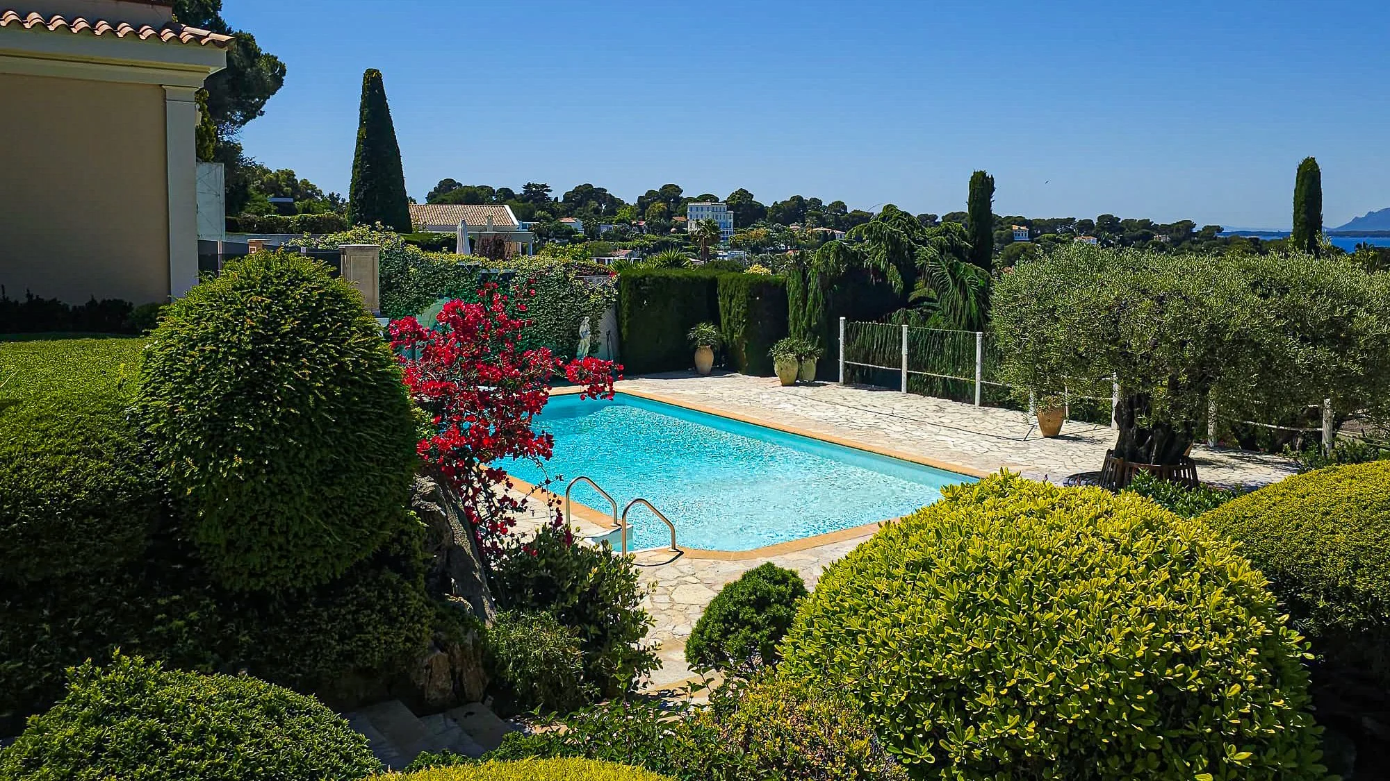 Piscine dans un jardin méditerranéen avec des arbustes verts, des arbres et des fleurs colorées, sous un ciel clair et bleu.