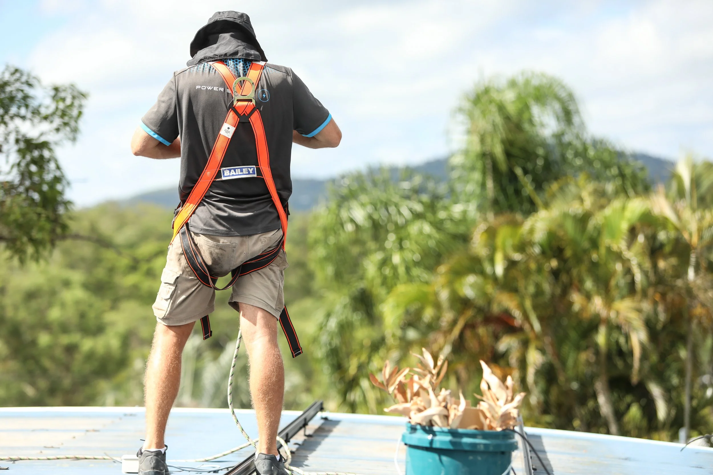 A person standing on a boat, facing away, wearing a black shirt with blue trim, beige shorts, and a safety harness. They are surrounded by lush green trees and blue sky in the background, with what appears to be a bucket with plant cuttings in front.