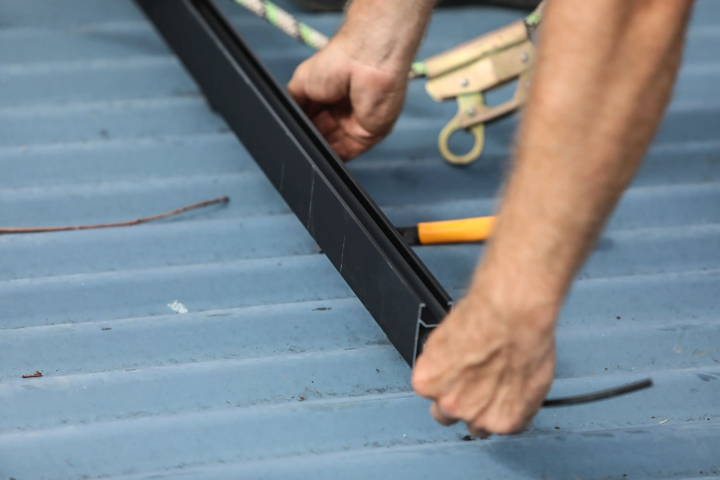 A person working on a metal gutter or similar structure, using a hand tool, with a ladder and a yellow-handled screwdriver visible on the corrugated metal surface.