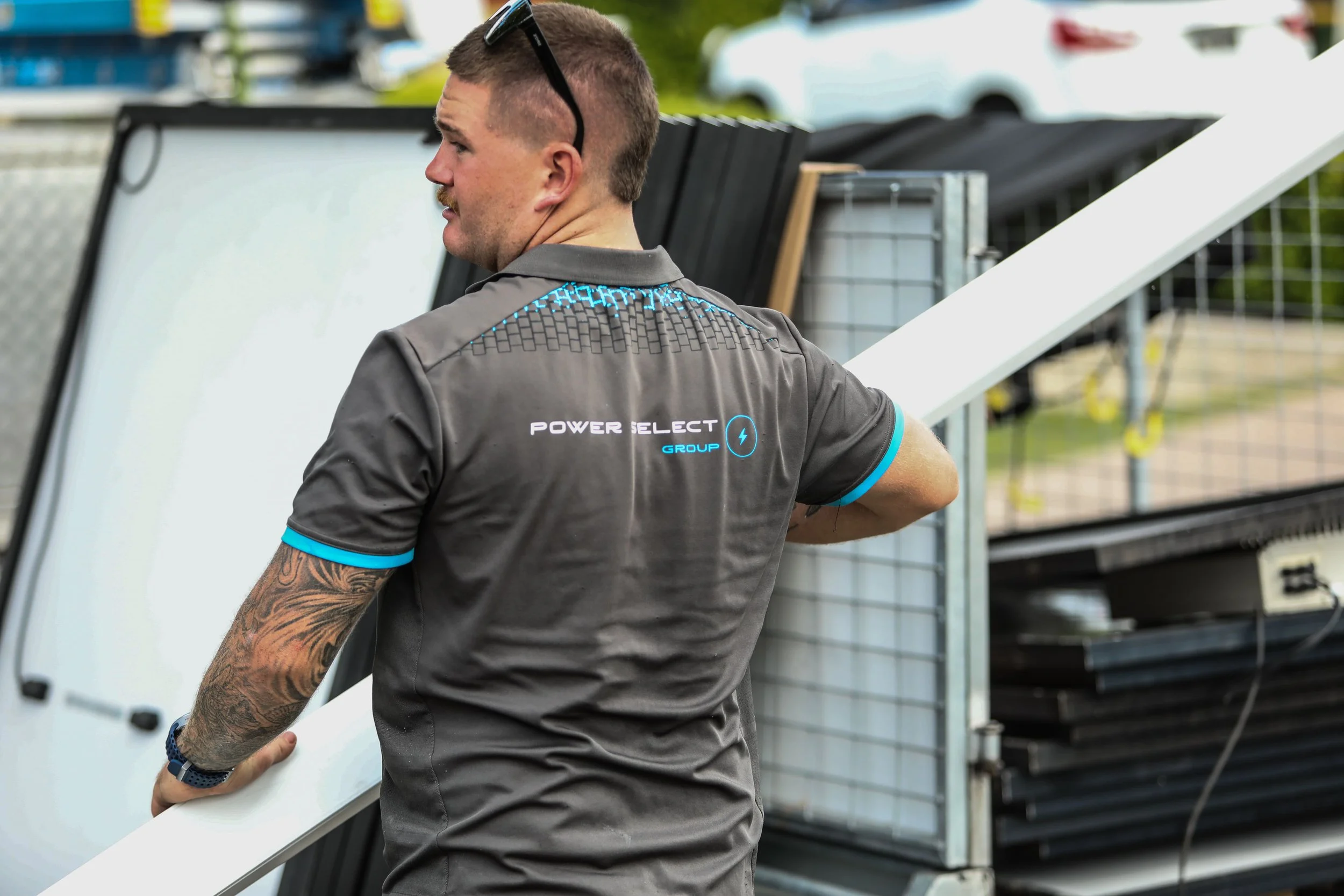 A man wearing a gray shirt with blue accents, standing outdoors, looking to the side, with sunglasses on his head, holding a large white panel. There are outdoor equipment and a white vehicle in the background.