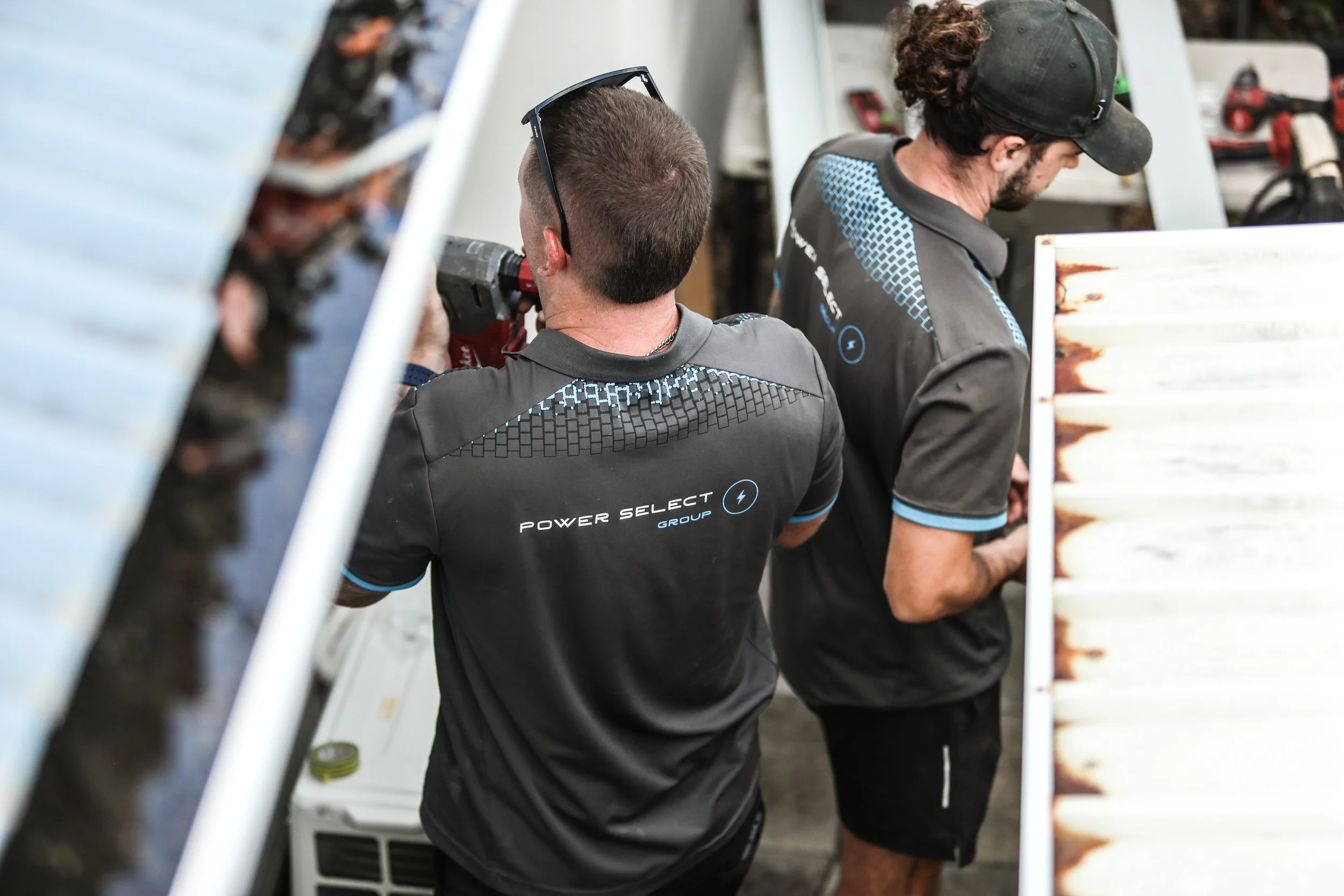 Two men wearing black and blue shirts labeled 'Power Select Group' working on equipment, one holding a power drill and the other looking at the equipment.