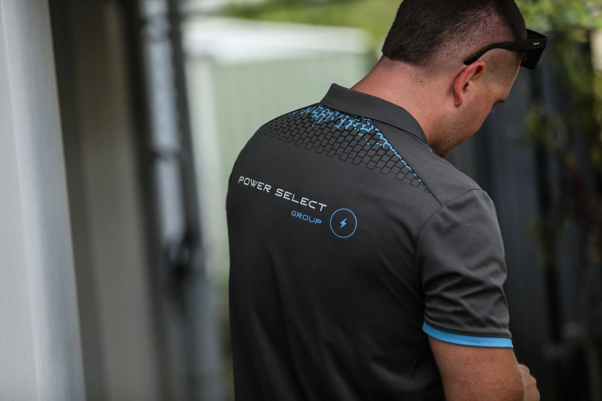 Man in gray shirt with black and blue details, wearing sunglasses, facing away, standing outdoors near a wall.