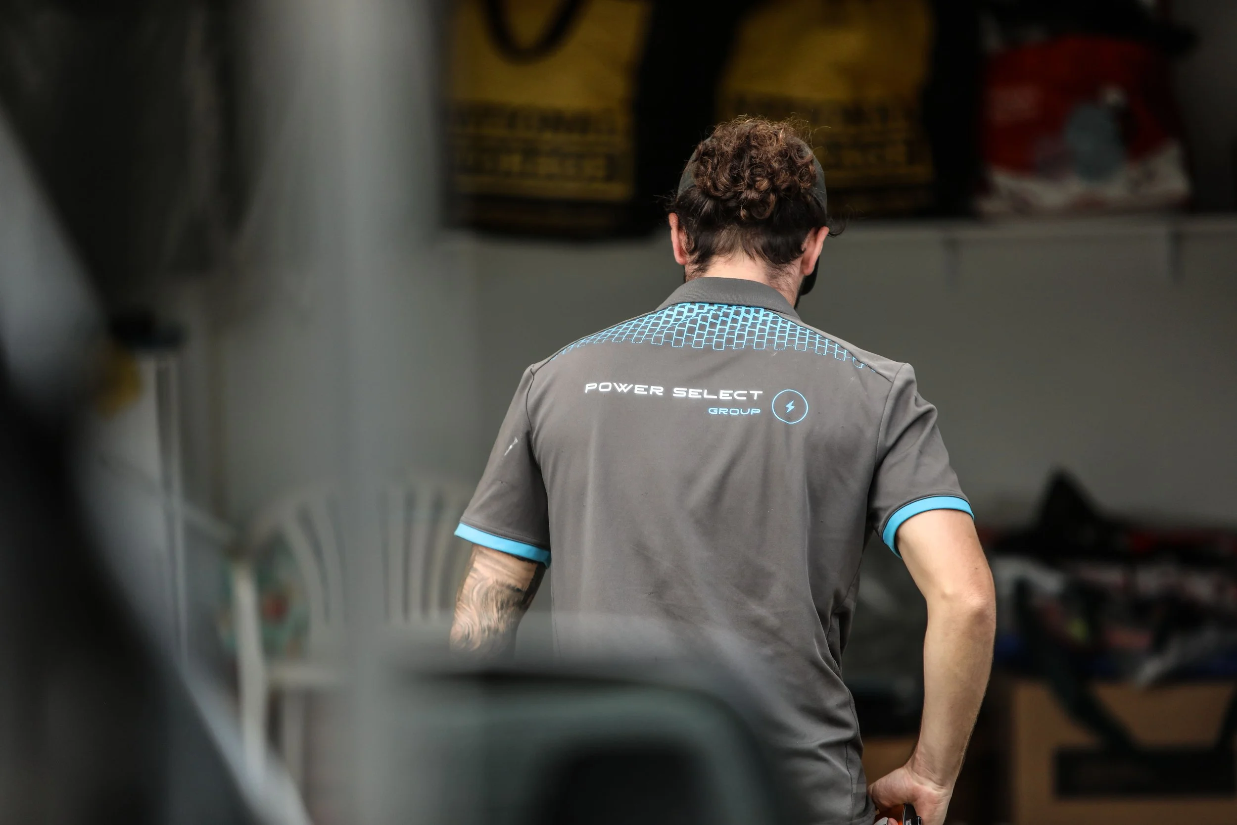 Back of a person wearing a gray shirt with blue accents and the text 'POWER SELECT GROUP' on the back, standing in what appears to be a workshop or storage area.