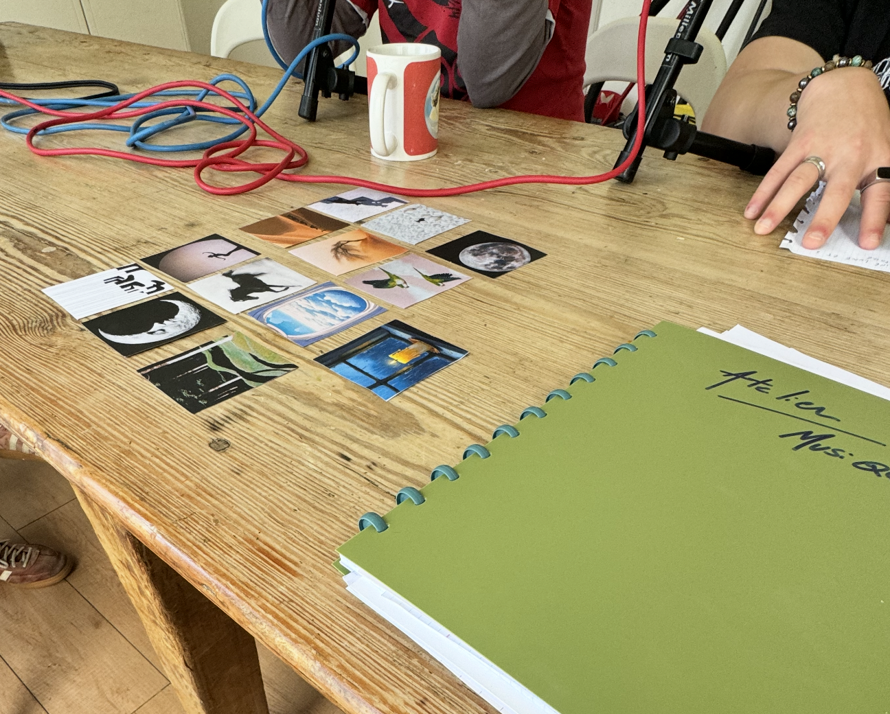 Table en bois avec des cartes illustrées disposées en ligne, une tasse, des microphones, des câbles rouges et bleus, un carnet vert et une main écrivant sur une feuille.