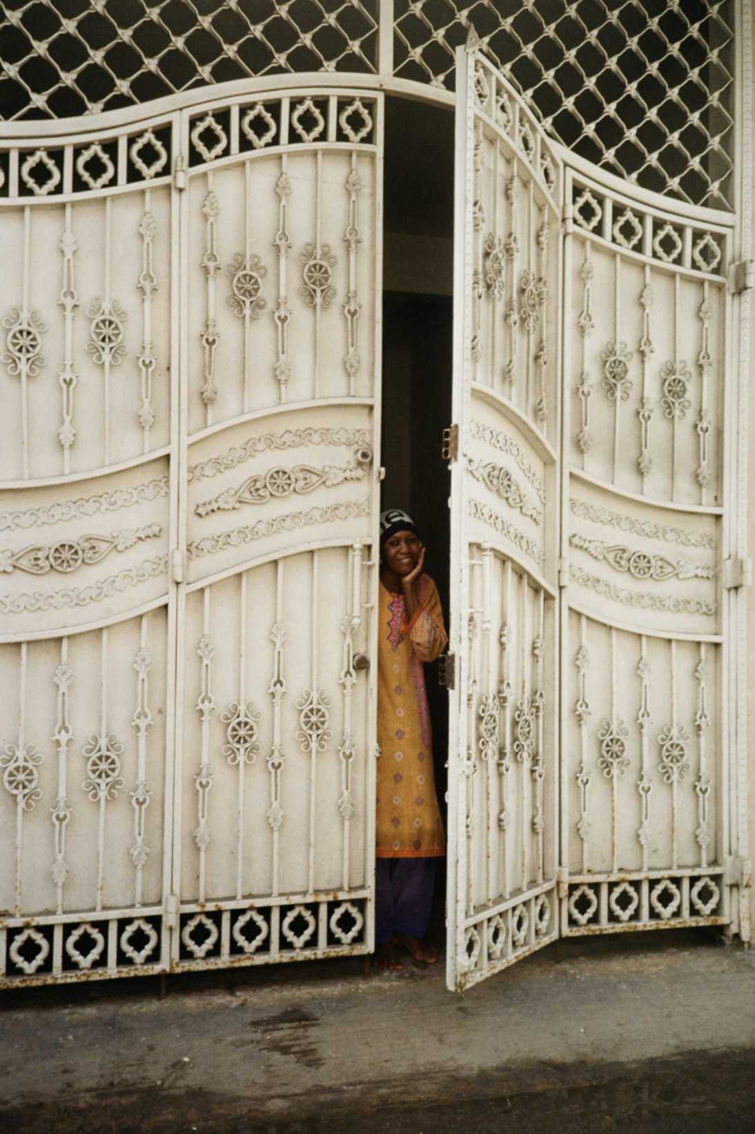 Une femme souriante, portant une robe jaune avec des motifs, se tient derrière une grande porte en fer orné de détails décoratifs, entrouverte.