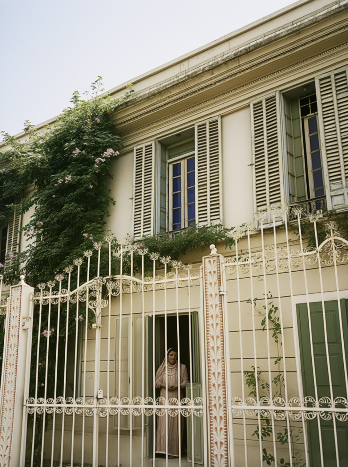 Une maison ancienne de couleur beige avec des volets vert et des fenêtres à barreaux, entourée d'une clôture en fer orné de détails décoratifs. Une femme habillée d'une robe beige se tient à la porte de la maison, derrière la clôture, avec des plante